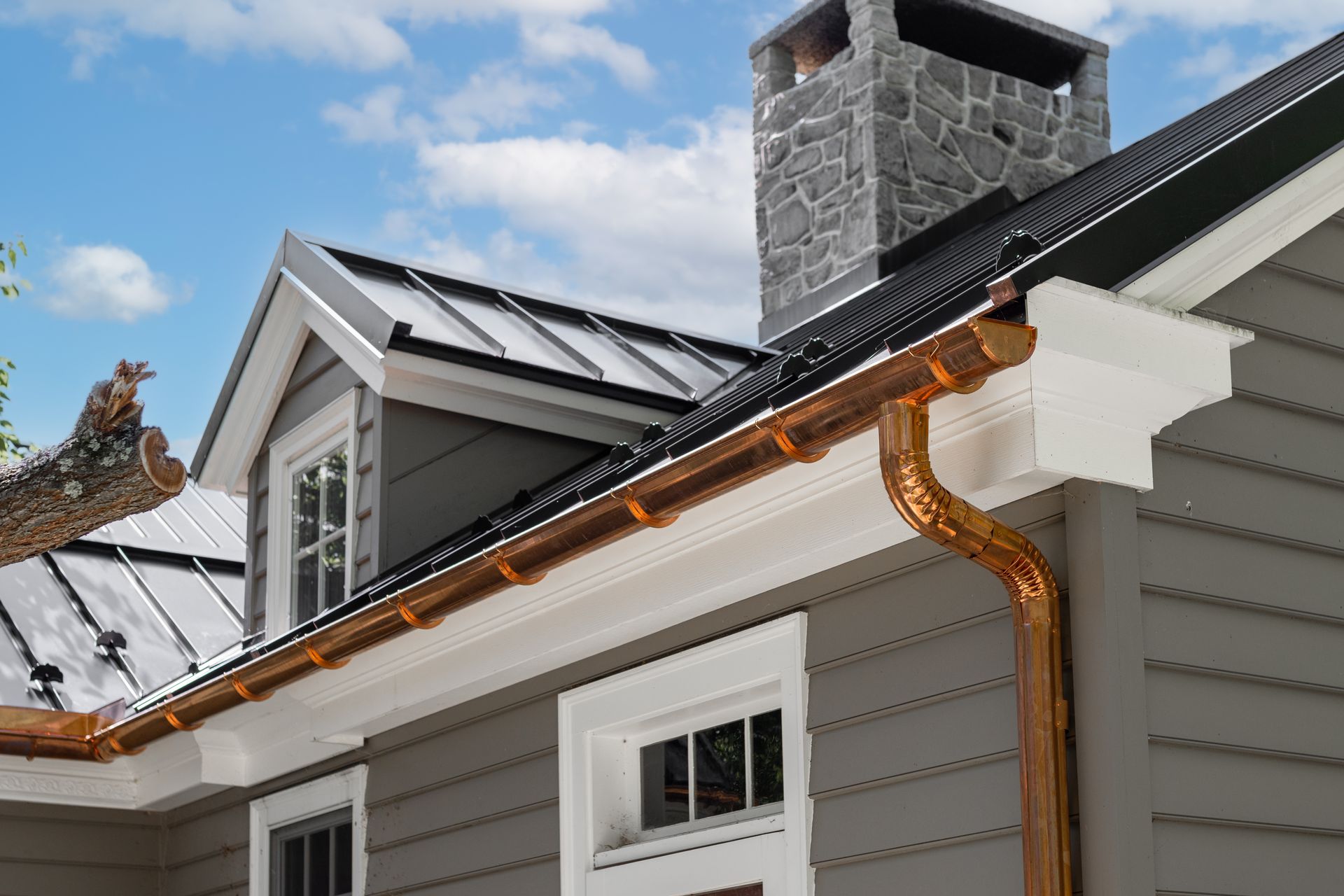 Copper gutters on a gray house with black roof and stone chimney against a blue sky.