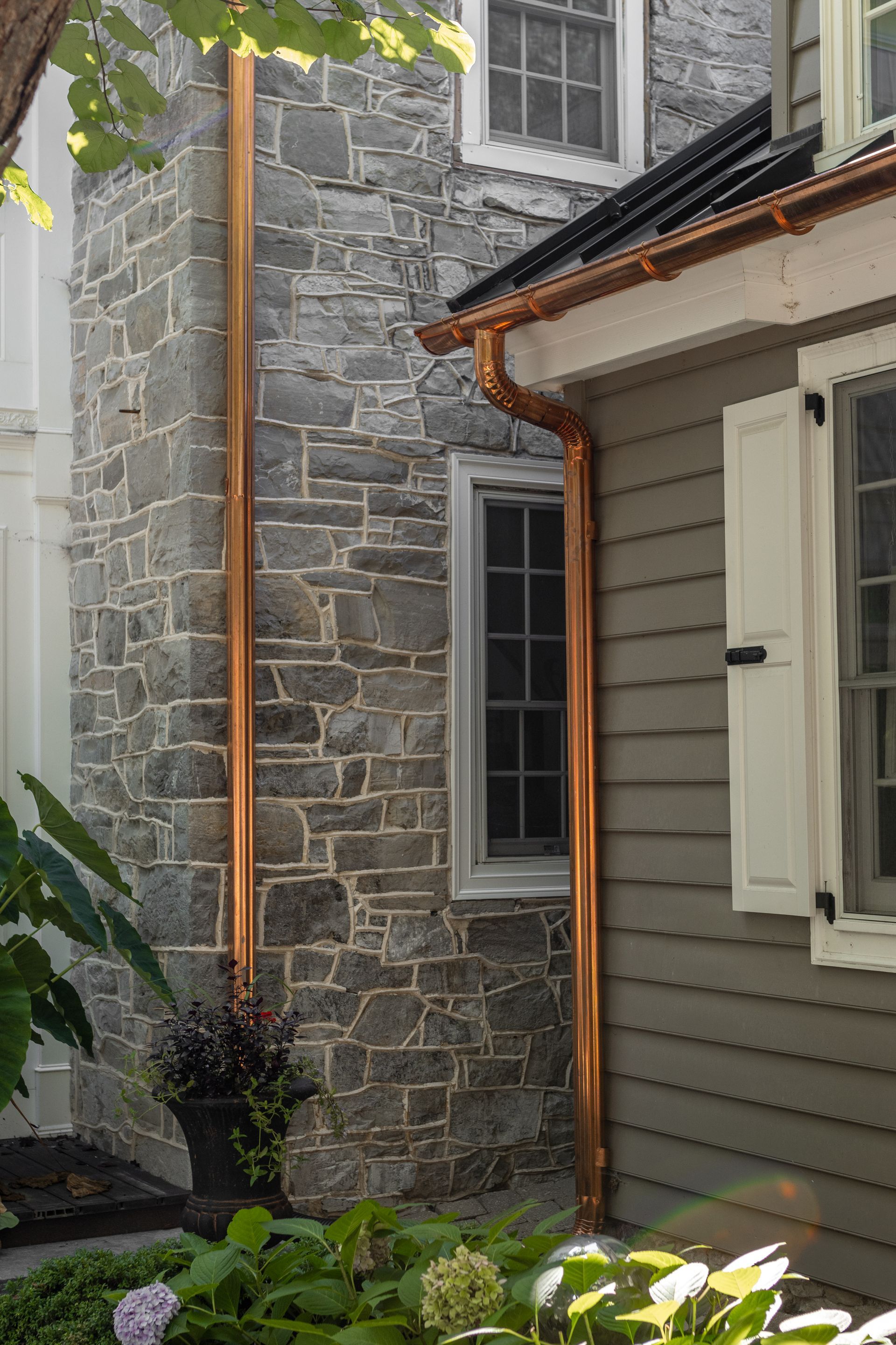 Copper rain gutters run along a stone wall and a gray-sided house with white shutters.