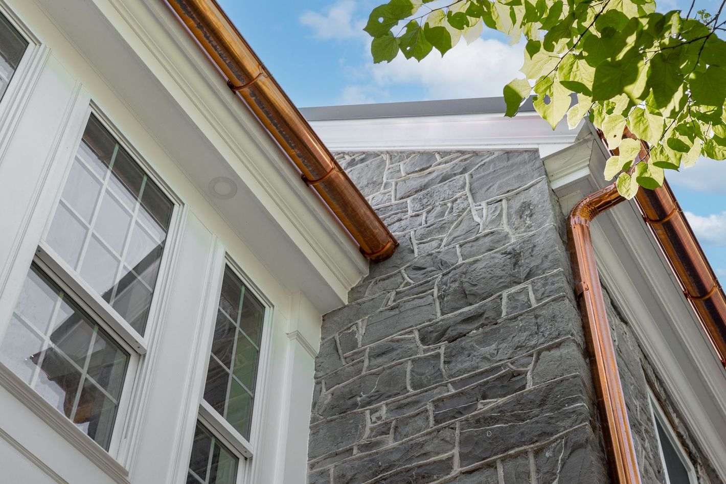 Copper gutters on building with stone wall and white trim. Blue sky and green leaves.