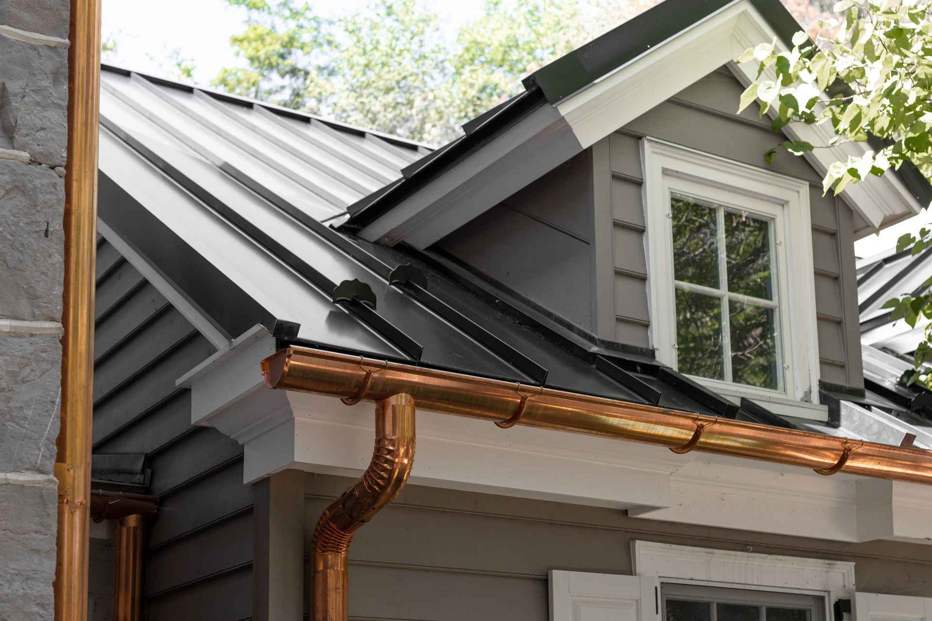 Black metal roof with copper gutters on a gray house with a dormer.