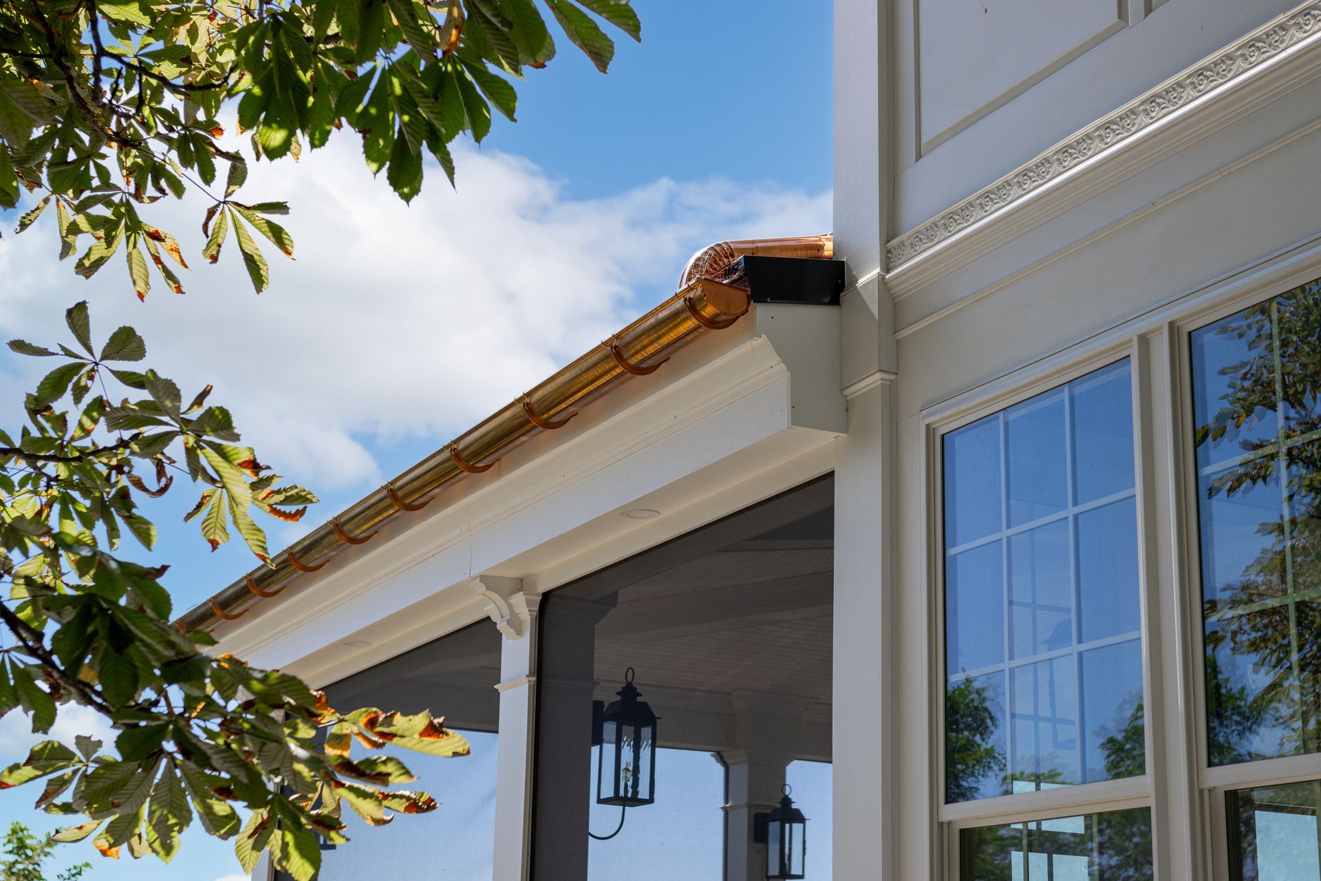Golden gutters on a white house with blue sky, some tree leaves, and screened porch visible.