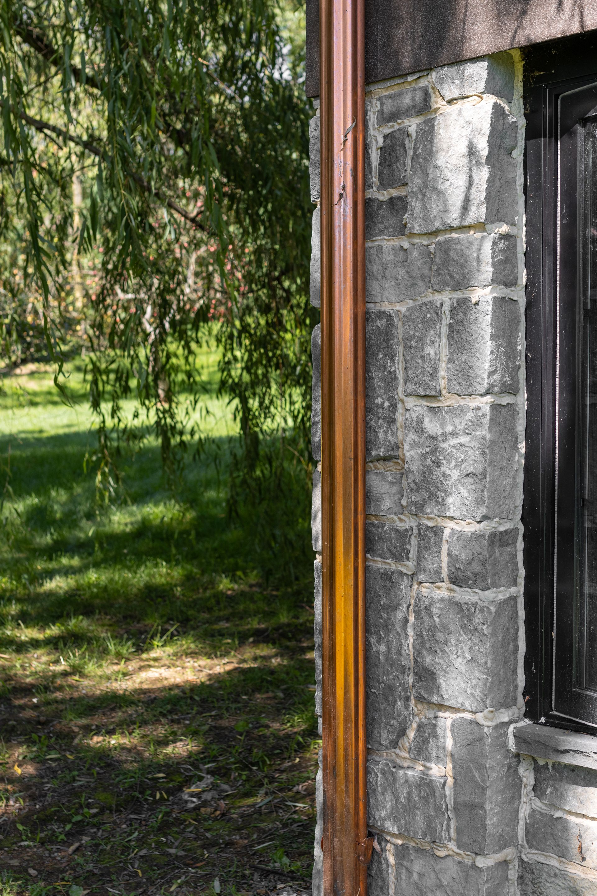 Copper downspout on a gray stone building exterior, with a window and green trees in the background.