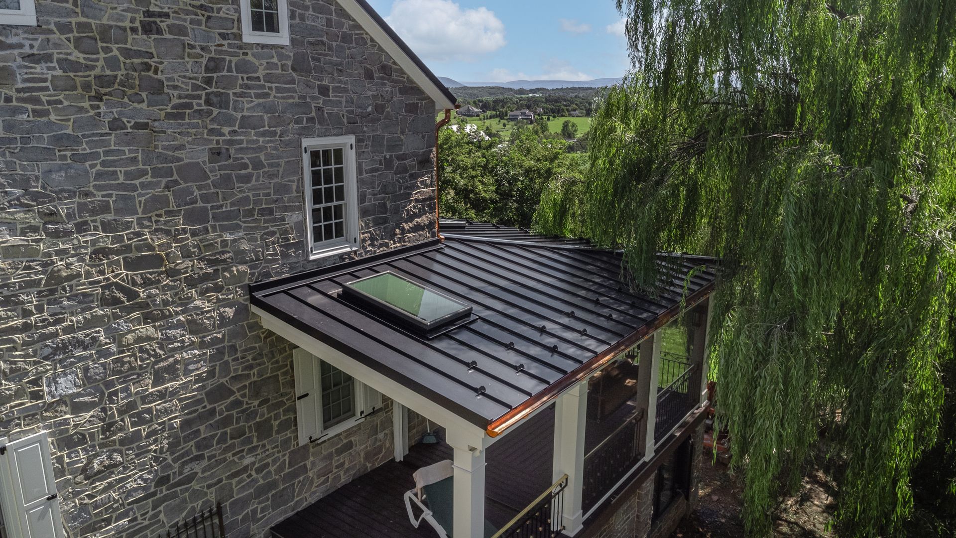 Stone house with a porch featuring a metal roof and skylight, with a weeping willow tree in the background.