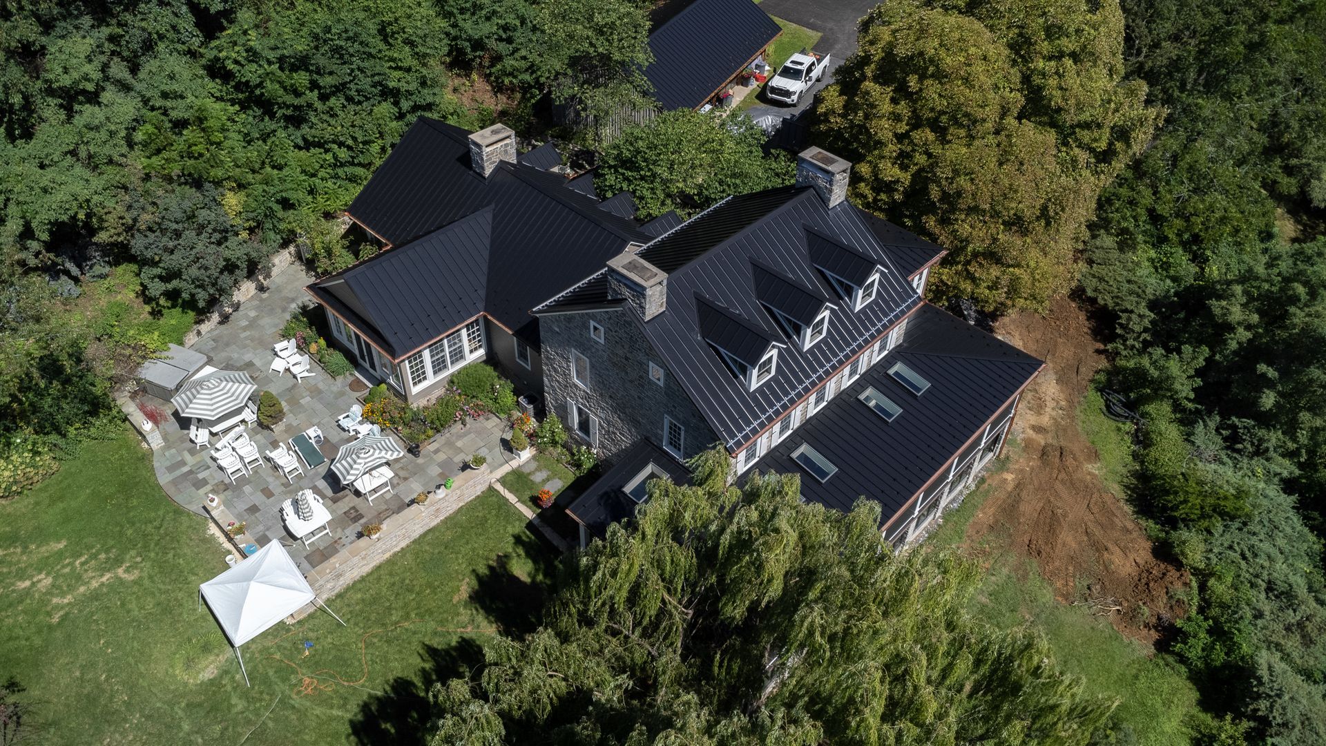 Aerial view of a large house with dark roof surrounded by trees and a patio with tables.