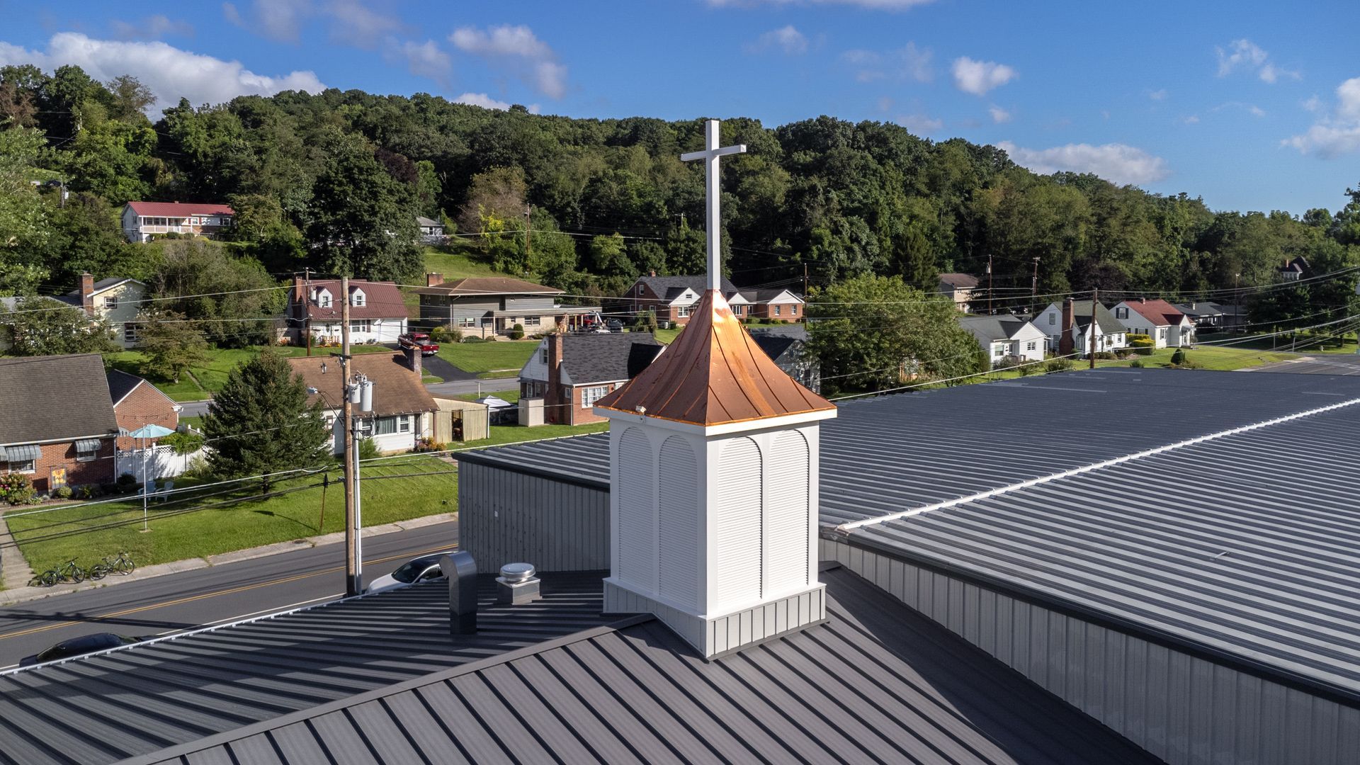 Rooftop steeple with cross; houses and wooded hill in the background.