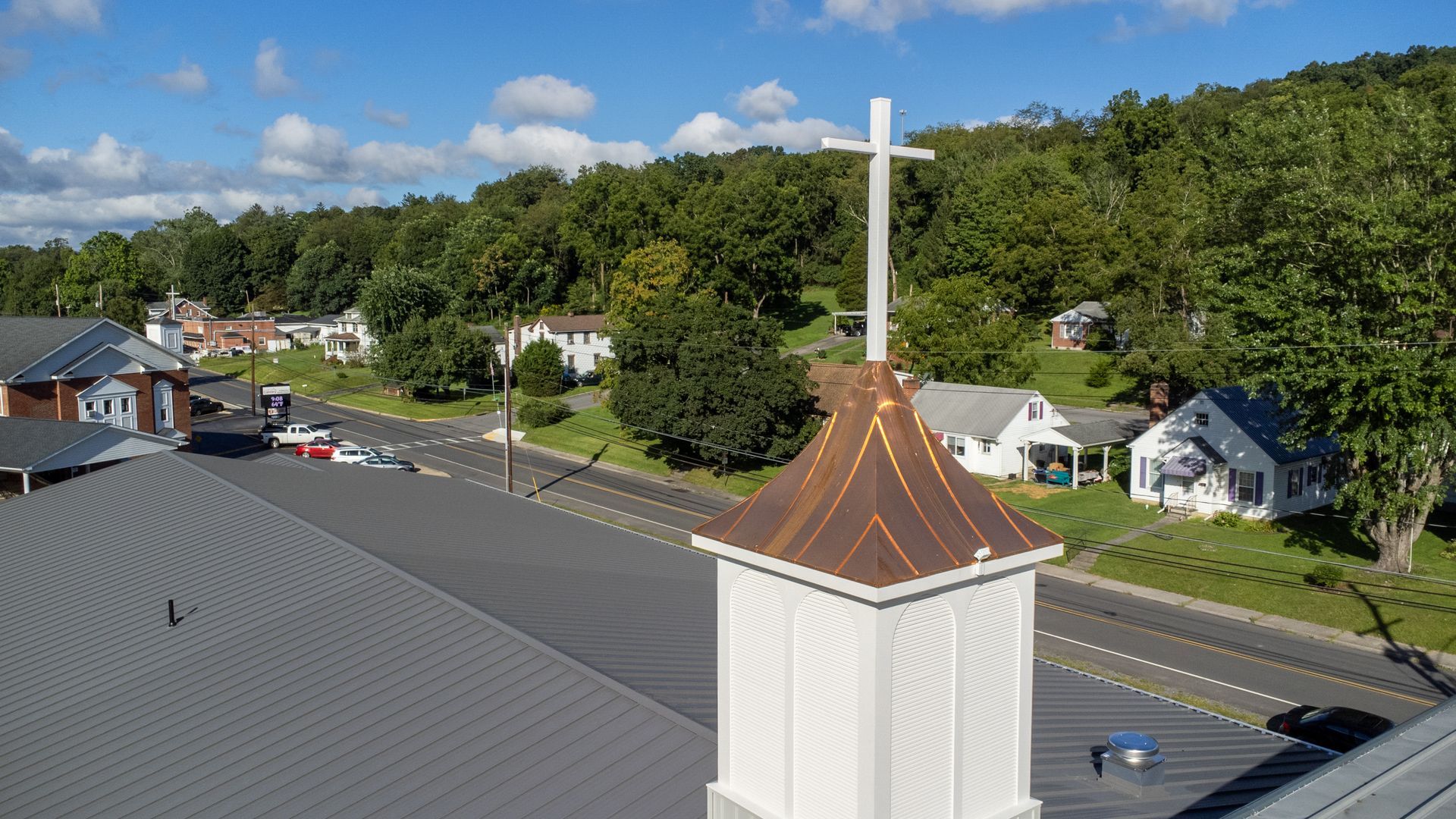 Church steeple with cross, overlooking a road with houses and a treeline under a blue sky.
