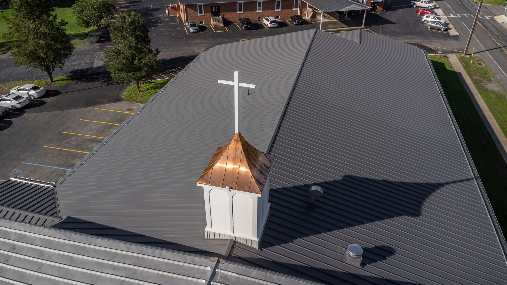 Aerial view of church with a copper-topped steeple, cross, and grey metal roof.