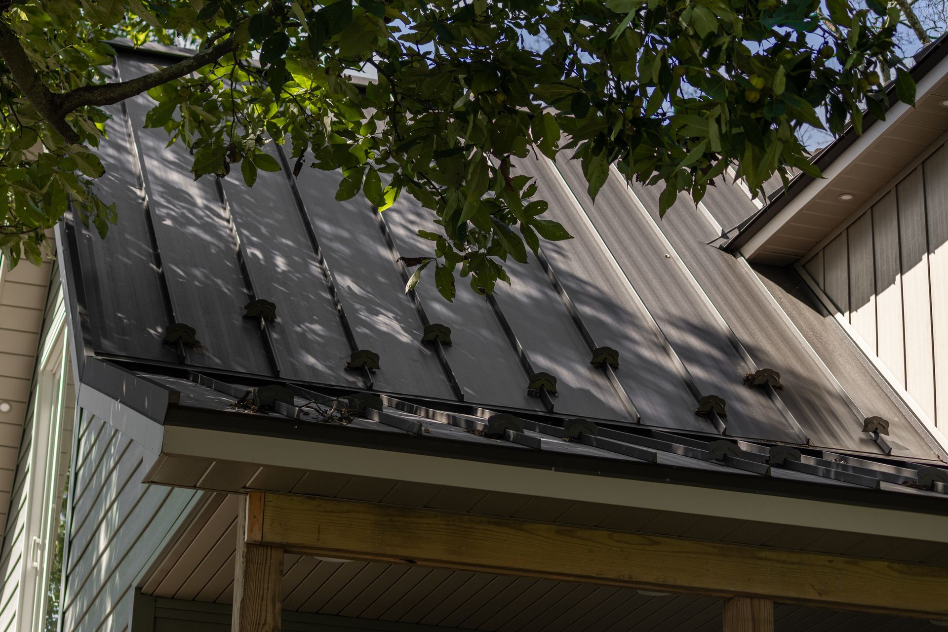 Black metal roof on a house, partially shaded by a tree, with a porch visible below.