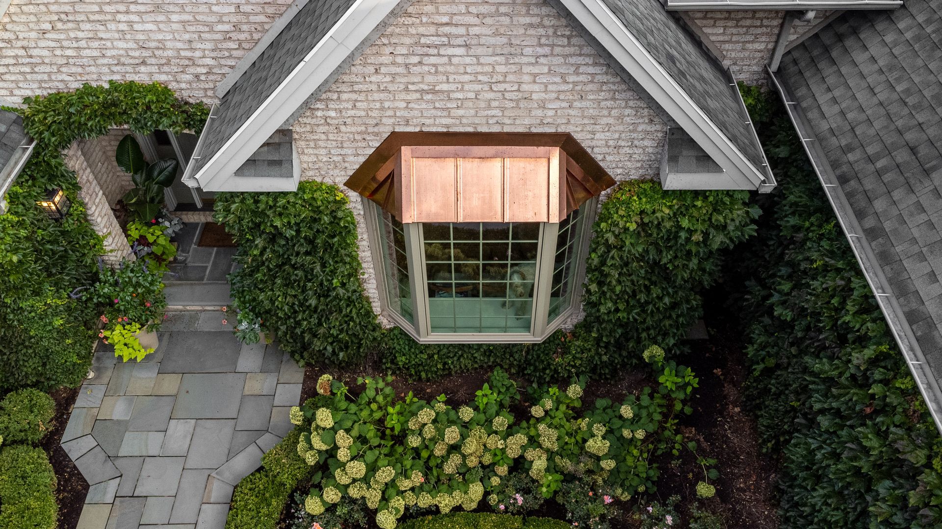 Overhead view of house with copper-roofed window, stone facade, slate walkway, and lush green landscaping.