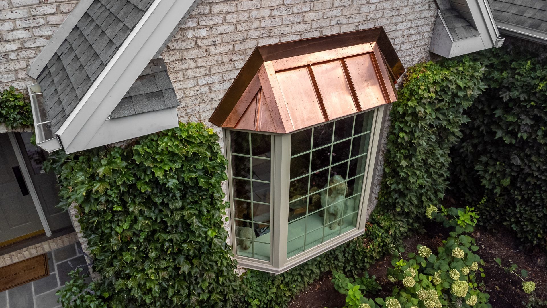 Copper-roofed bay window on a brick house covered in ivy, with the gray roof of the building visible.