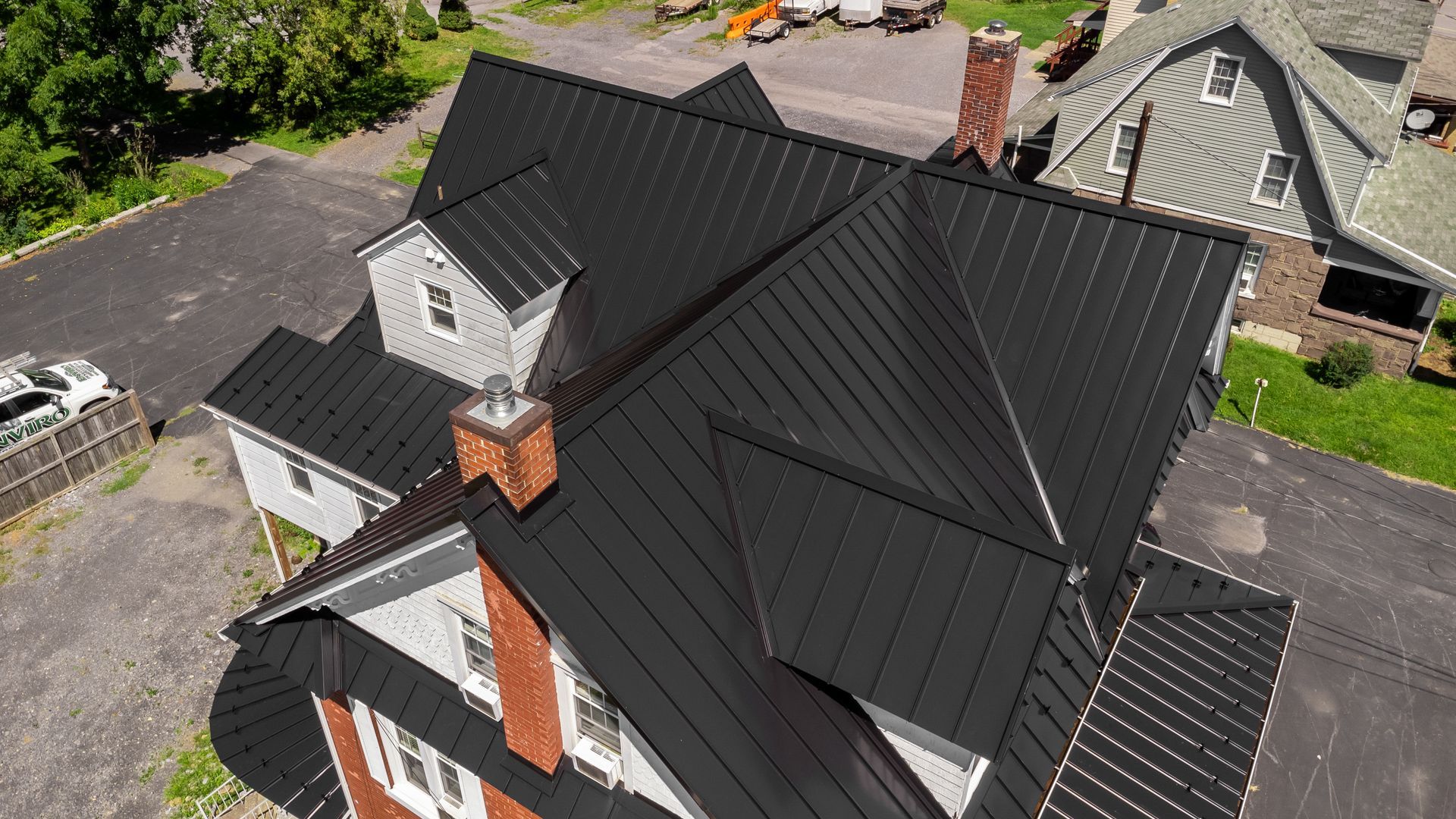 Black metal roof on a two-story house with a brick chimney and dormer. Sunny outdoor setting.