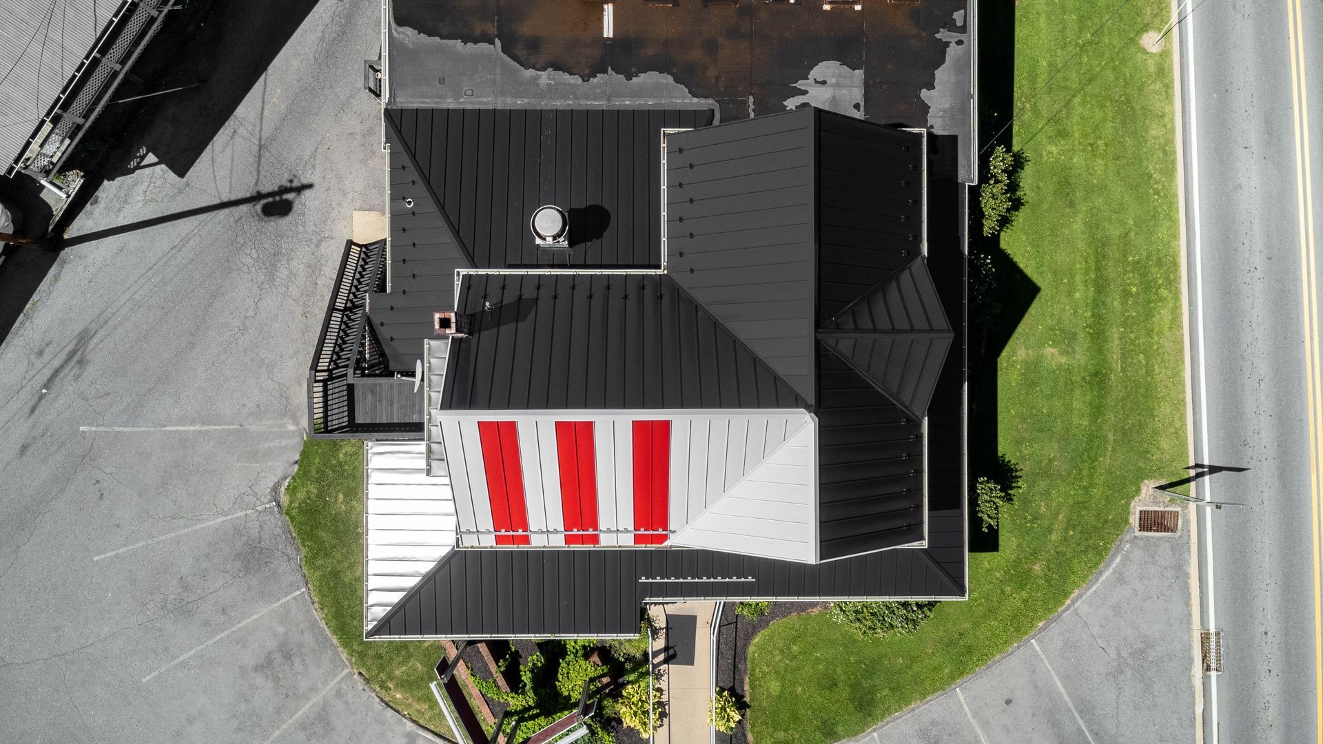 Aerial view of a building with a black and red roof. Building is next to grass and road.