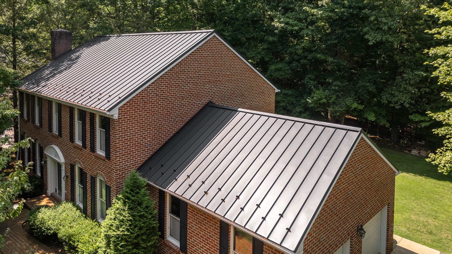 Brick house with dark metal roof surrounded by green trees.