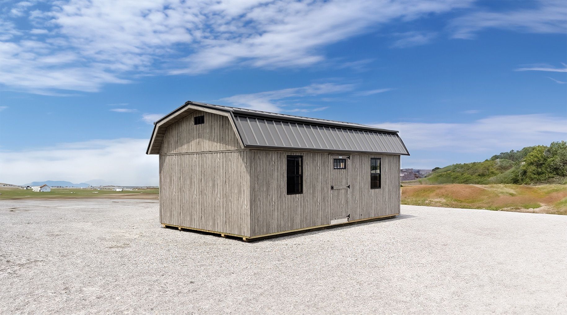 Barn-style shed with weathered wood siding, metal roof, and two windows, set on a gravel surface under a blue sky.