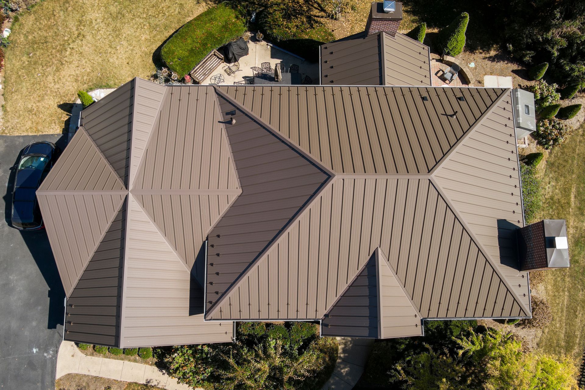 An overhead aerial view of a house with a metal roof with brown, patterned shingles, surrounded by a lawn and mature trees.