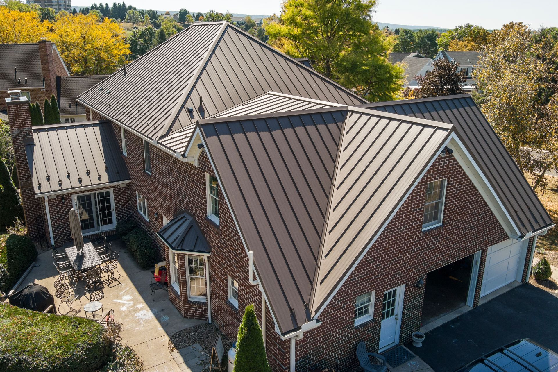 A brick house with a dark metal roof, featuring multiple gables, a garage, and a patio area on a sunny day.