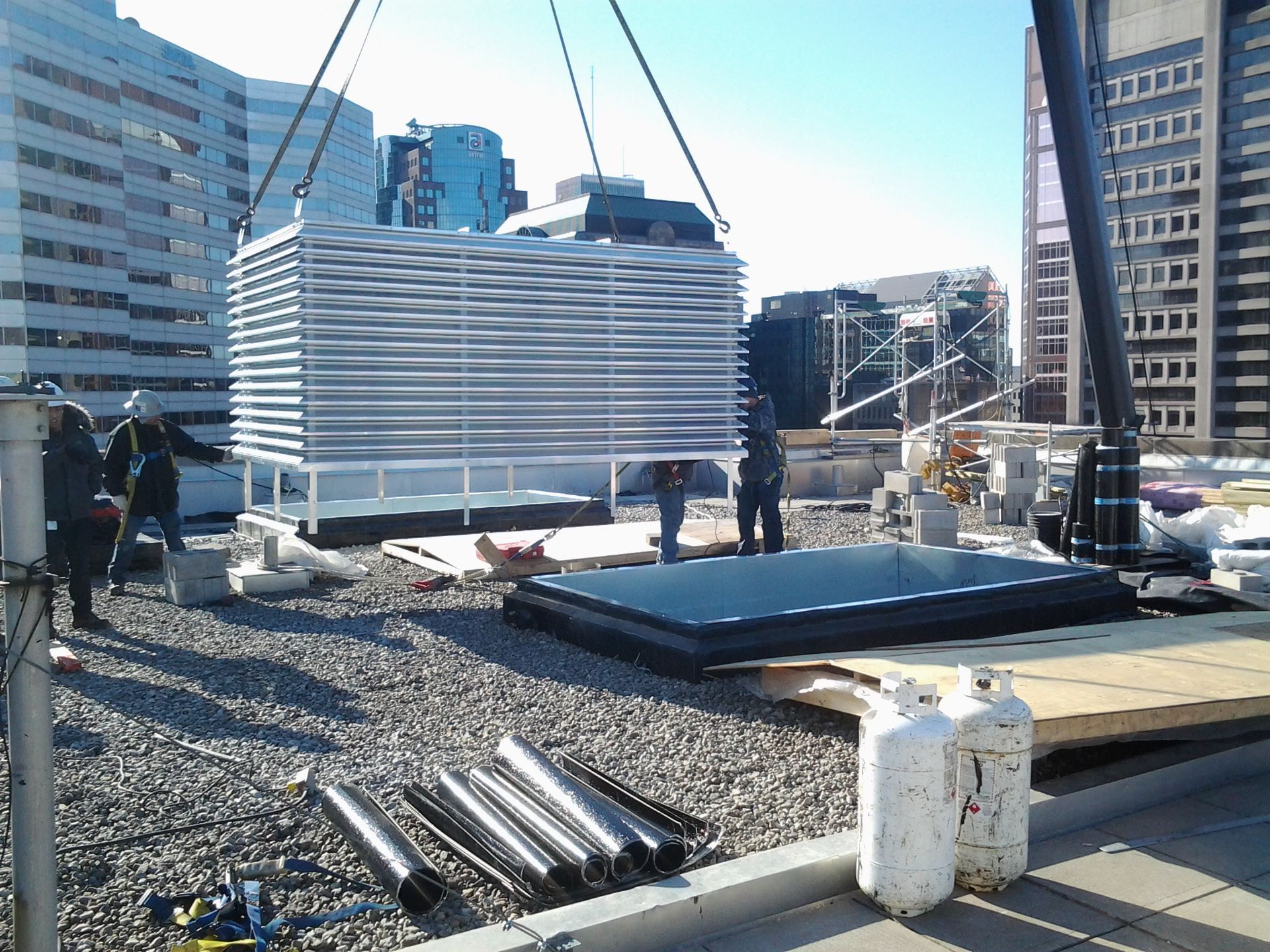 Une imposante unité de climatisation est hissée par une grue sur le toit d'un immeuble. Des ouvriers se tiennent à proximité.