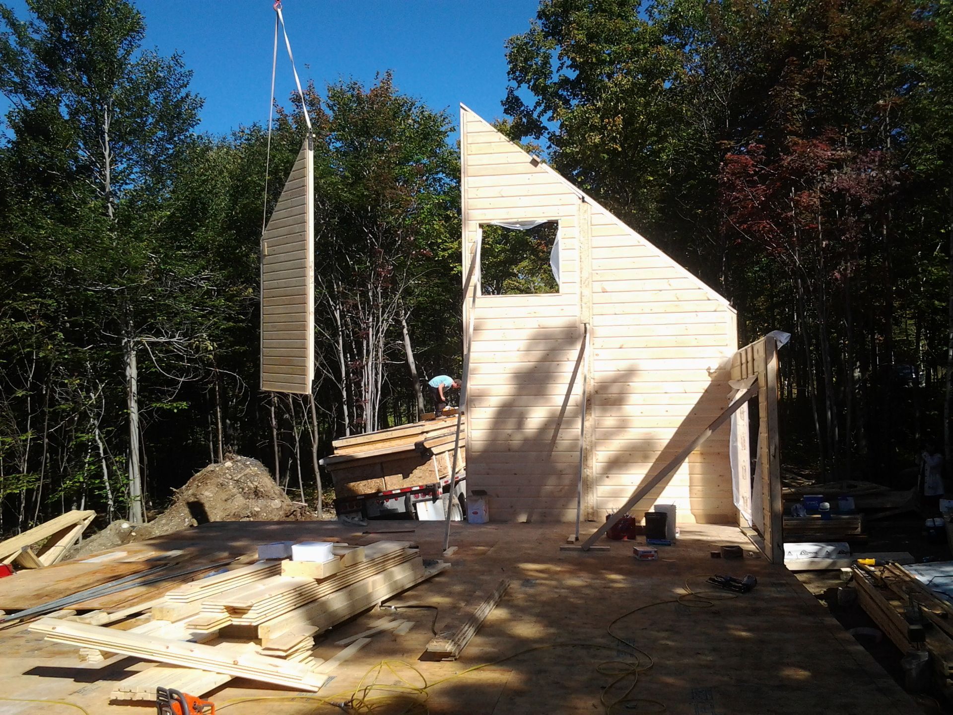 Chantier de construction : un panneau mural en bois est mis en place par une grue. Ciel bleu, forêt en arrière-plan.