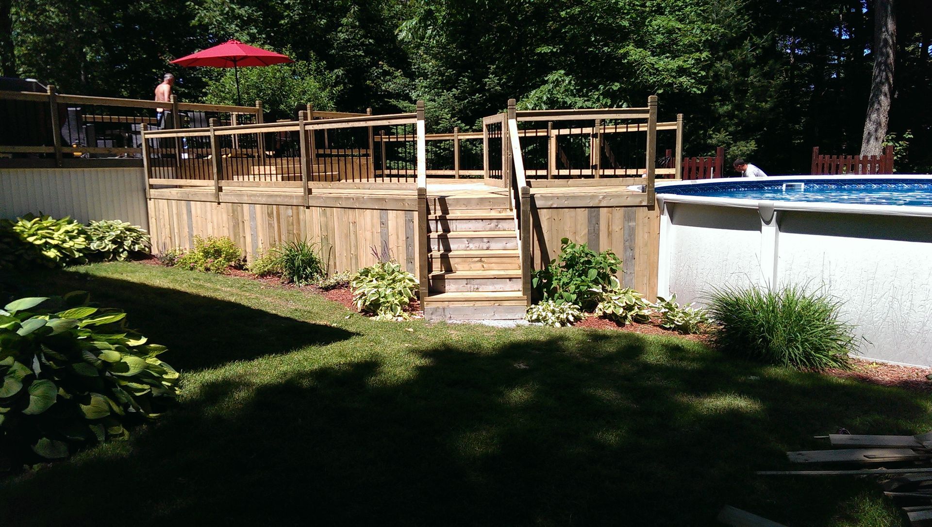 Terrasse en bois avec marches jouxtant une piscine et un aménagement paysager de jardin, parasol rouge visible.