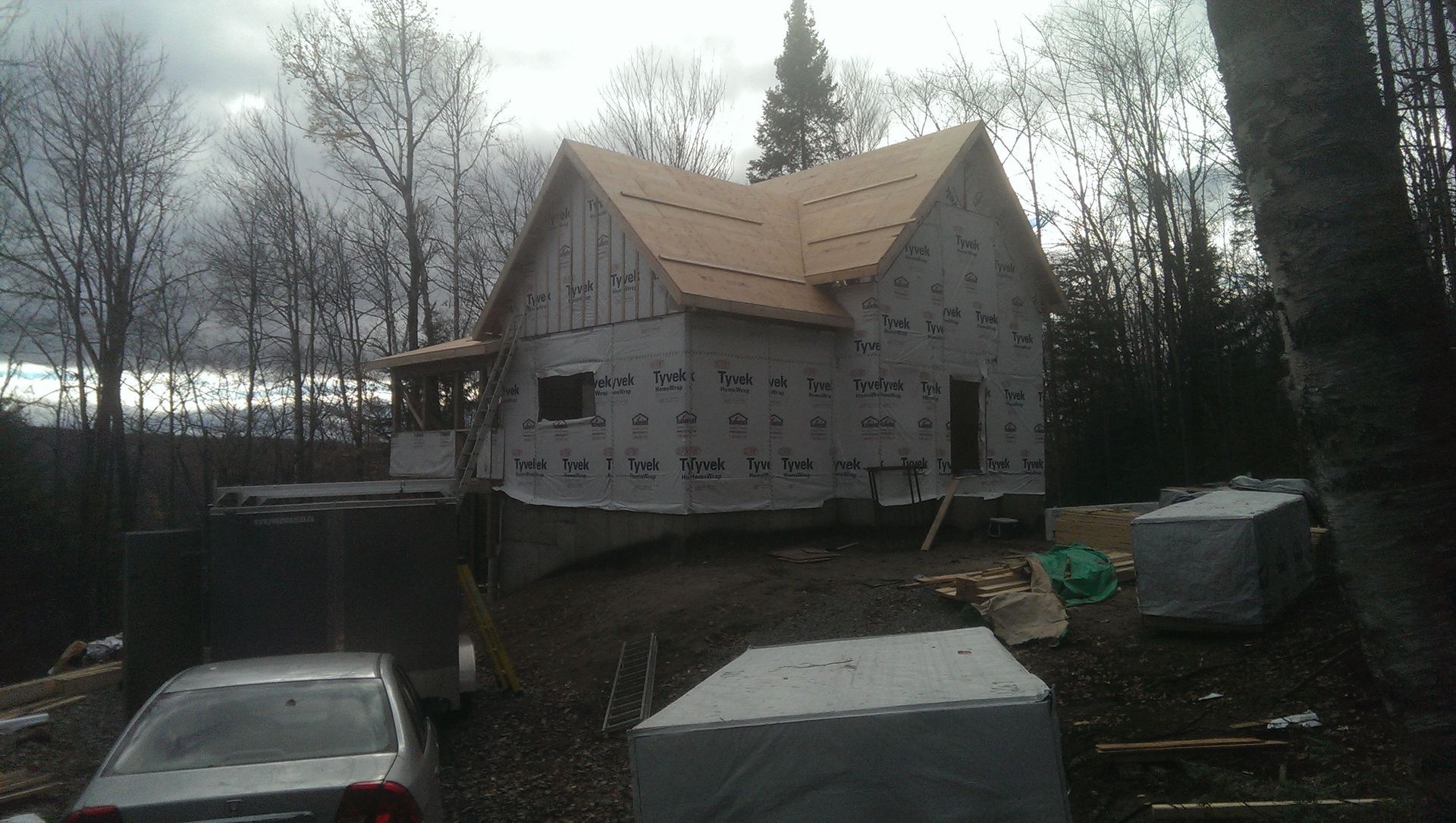 Maison en construction, bardage inachevé, toiture neuve, entourée d'arbres et de matériaux de construction.