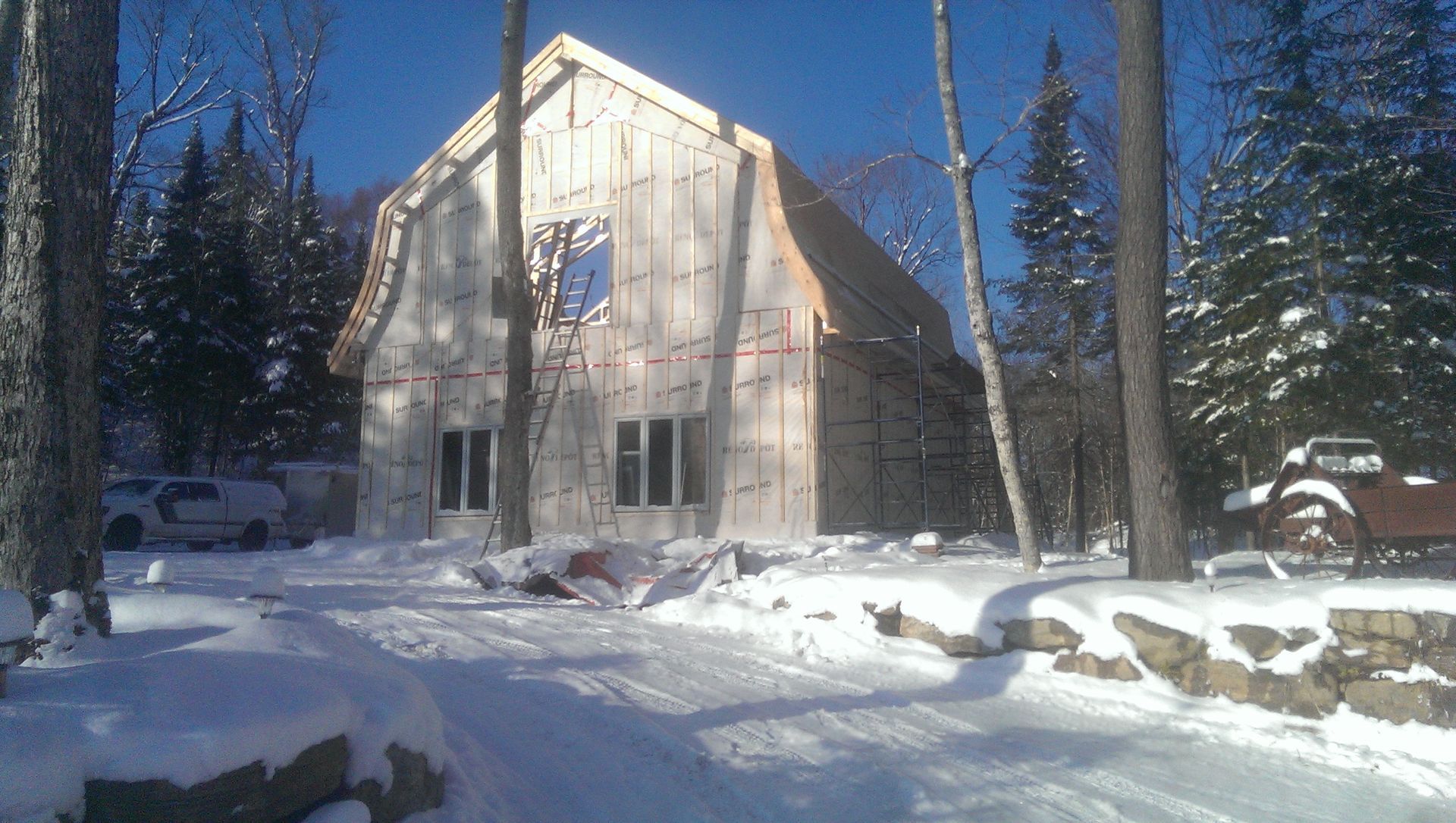 Chantier enneigé : structure de type grange entourée d’arbres ; ciel bleu.
