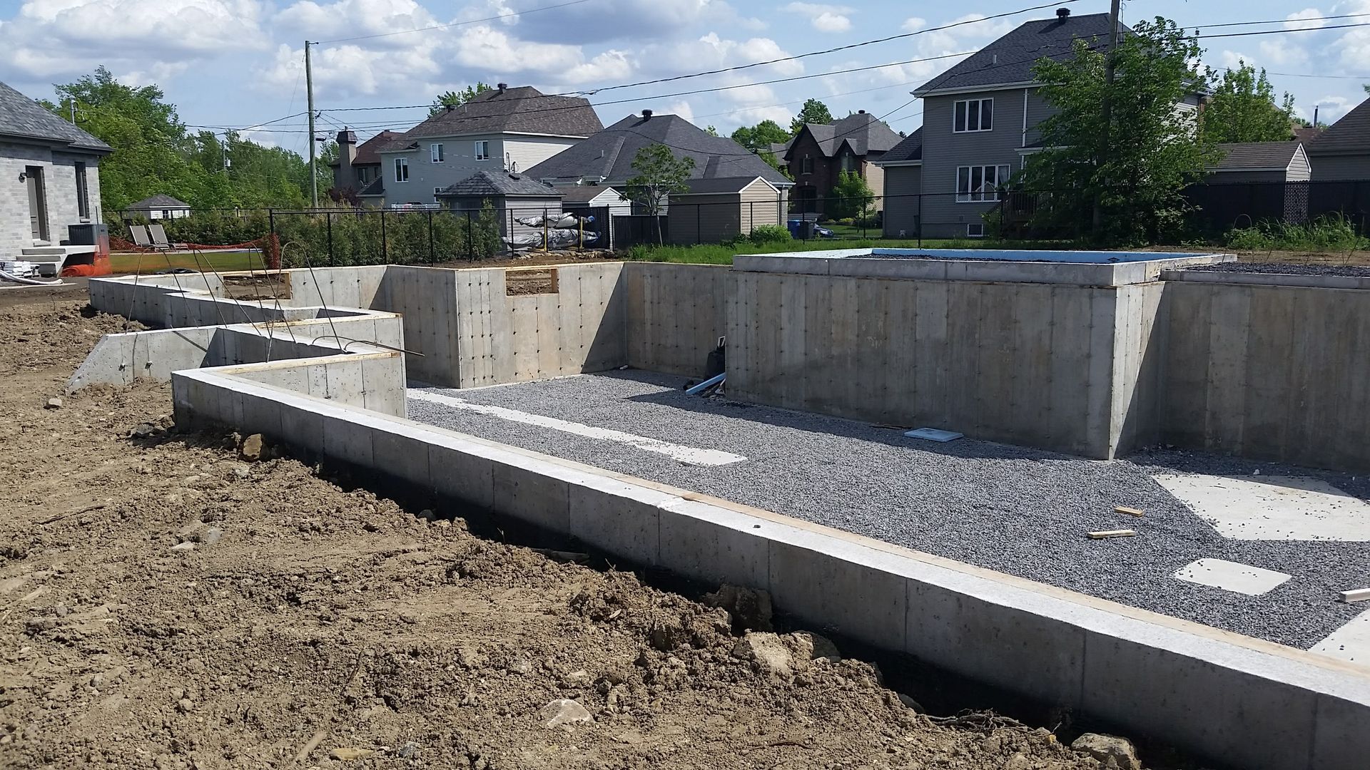 Fondations en béton pour une maison en construction ; intérieur rempli de gravier ; terrain vague environnant, avec des maisons en arrière-plan.