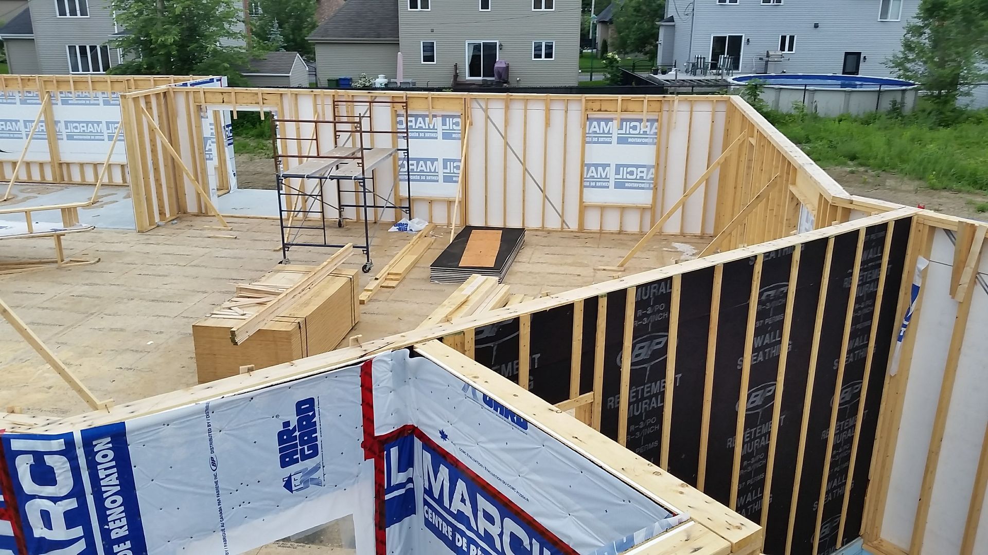 Ossature bois d'une maison en construction ; membrane d'étanchéité bleue sur les murs extérieurs, matériaux en bois éparpillés.