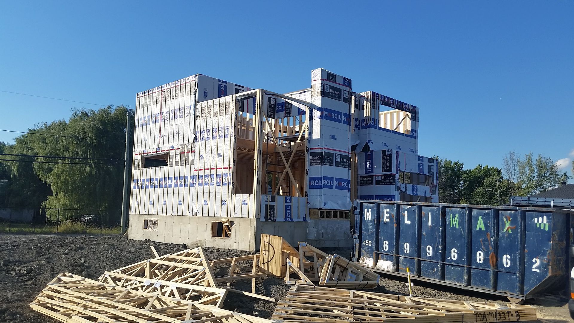 Bâtiment en construction, charpente en bois et bâche bleue, ciel bleu et bennes à ordures en arrière-plan.