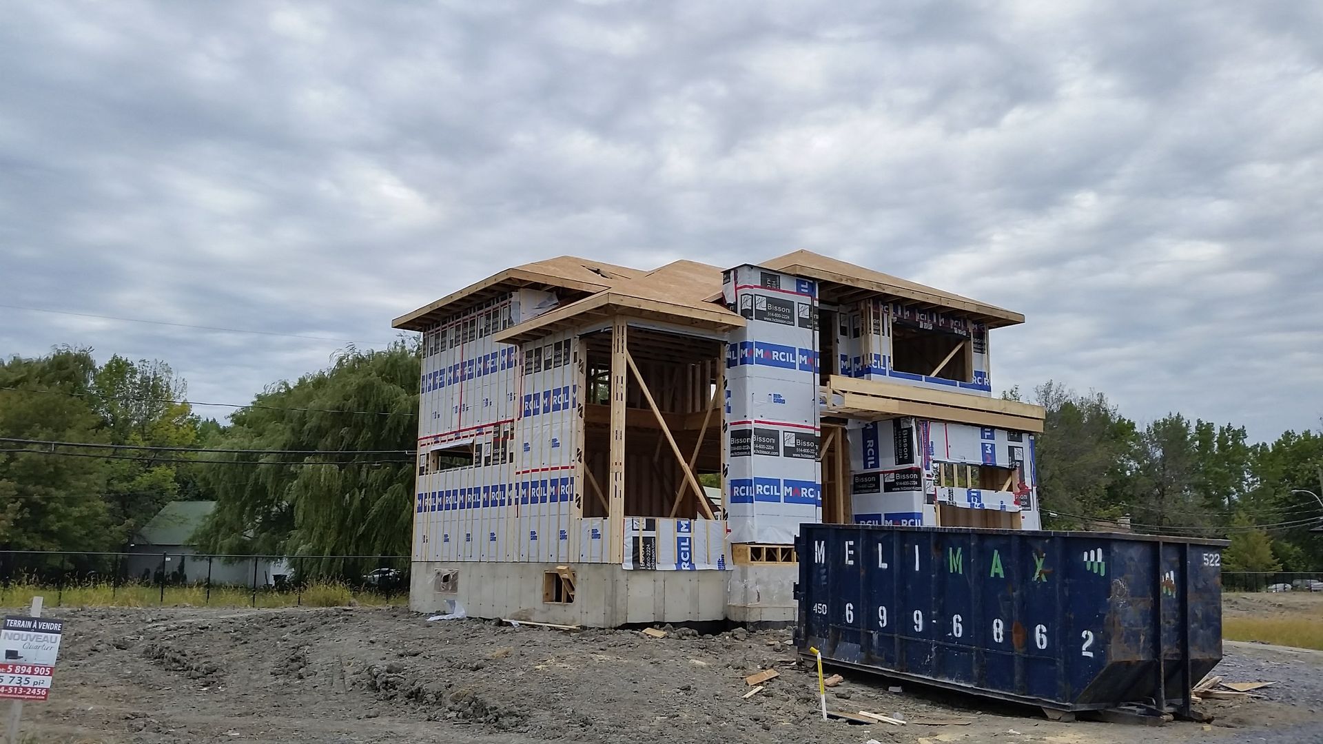 Maison à deux étages en construction, avec une ossature en bois et un revêtement bleu, à côté d'une grande benne à ordures.