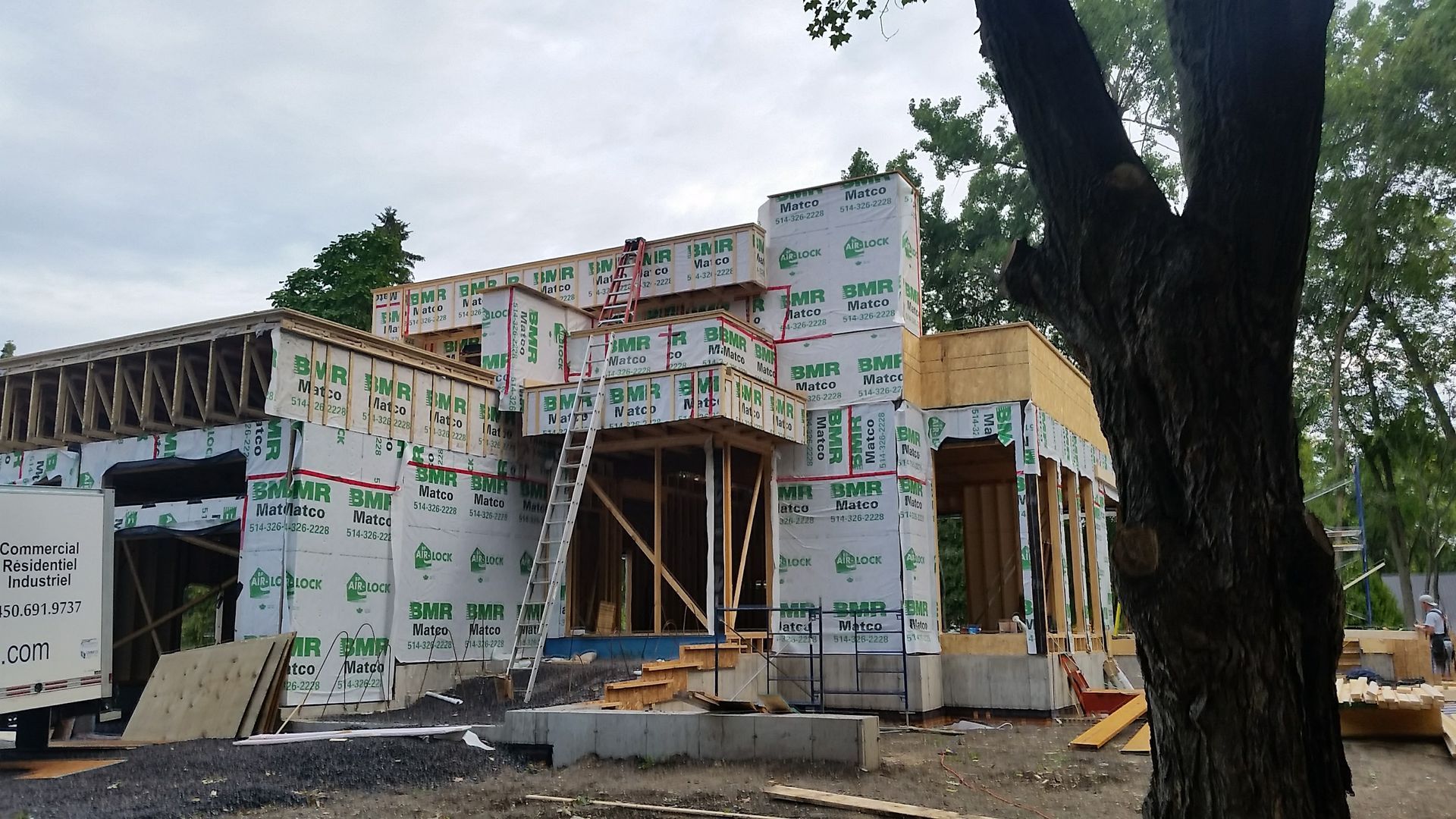 Maison en construction, ossature bois et bardage vert. Échelle menant au toit, arbres en arrière-plan.