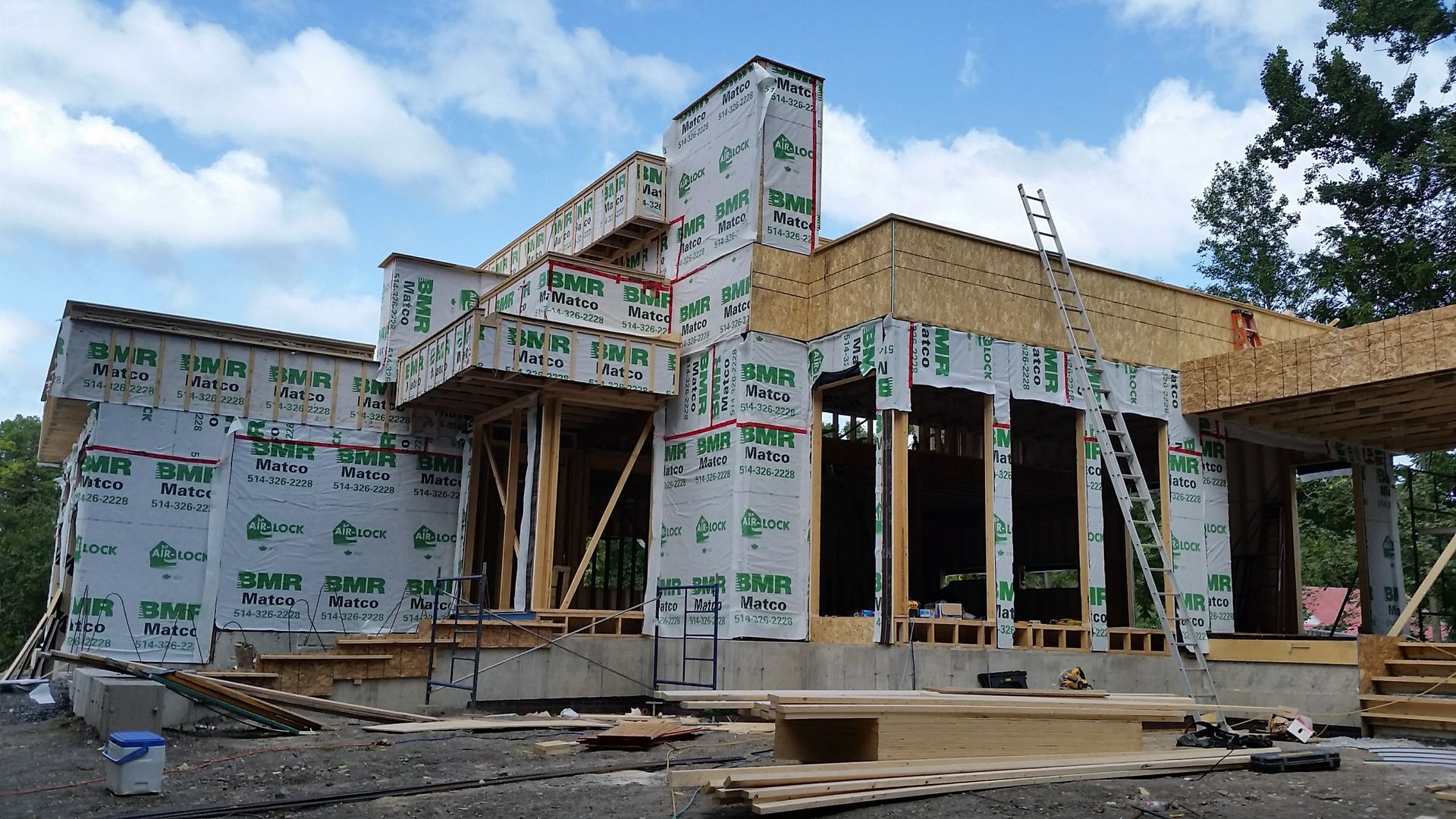 Maison en construction, vue extérieure. Charpente en bois recouverte d'un matériau de protection, sur fond de ciel nuageux.