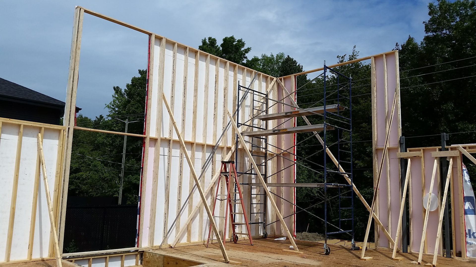 Ossature des murs extérieurs d'un bâtiment en construction ; poutres en bois, isolation blanche, échafaudages et arbres en arrière-plan.