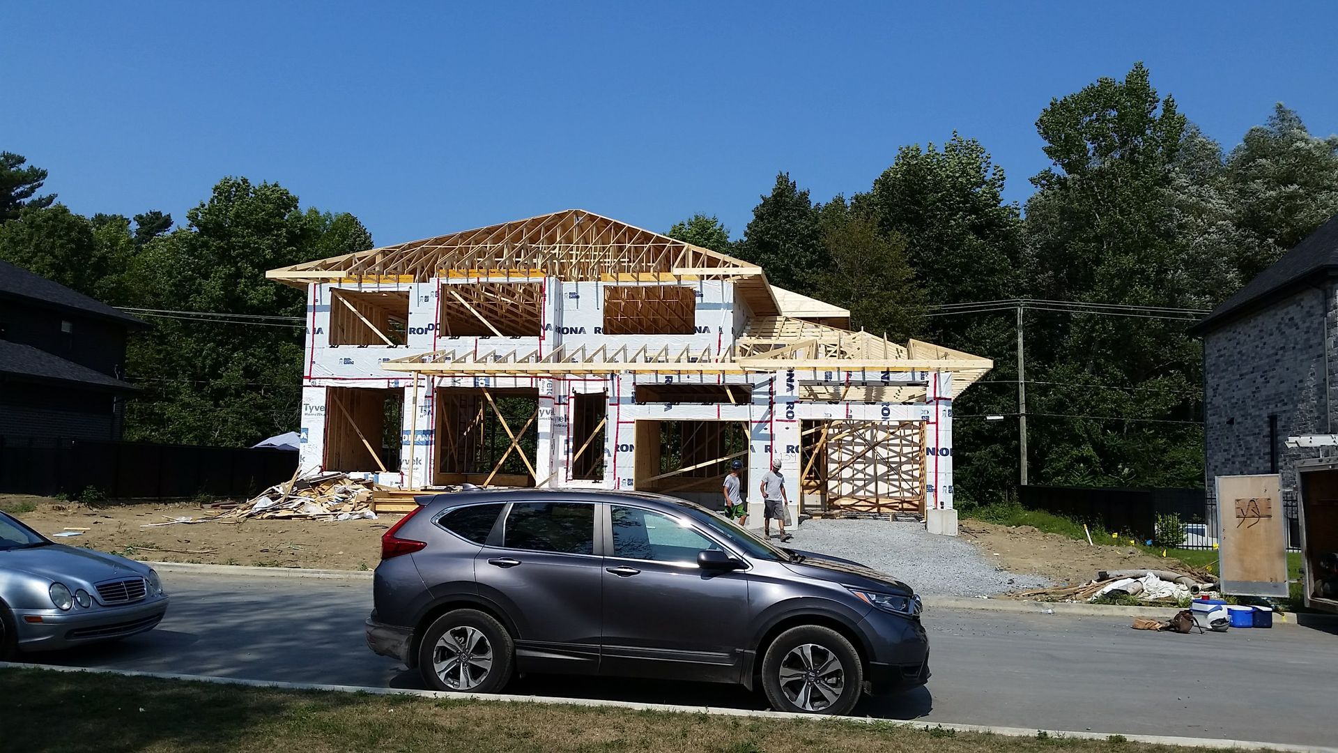 Maison en construction avec charpente apparente et voiture devant. Ciel bleu et arbres en arrière-plan.