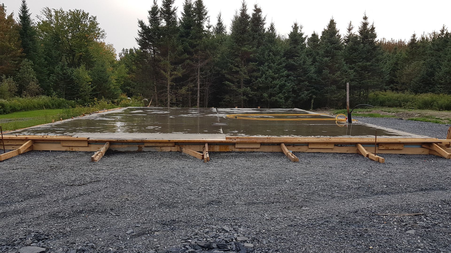 Un chantier de construction rempli de gravier, avec des coffrages en bois et de l'eau stagnante, la forêt en arrière-plan.