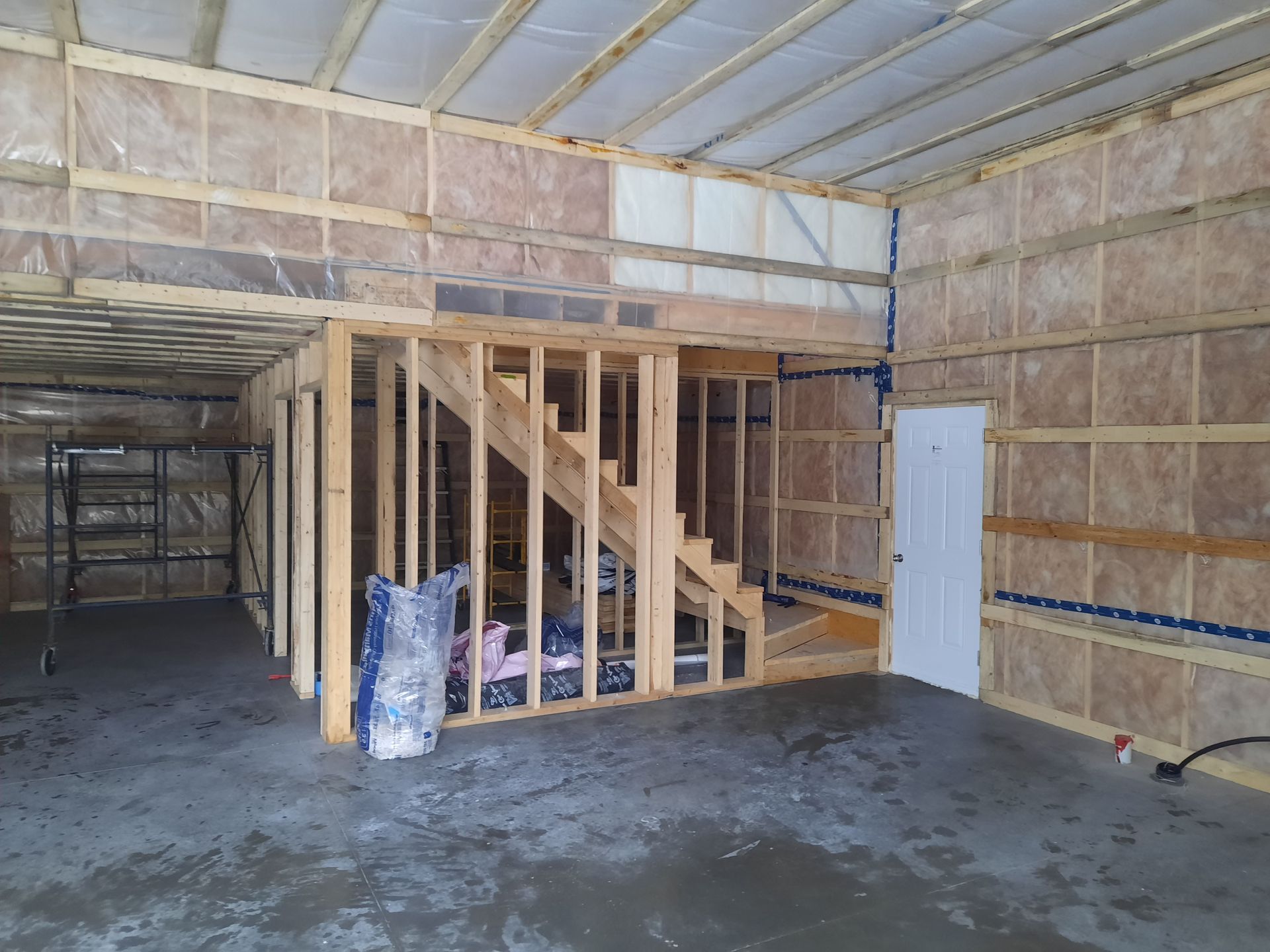 Intérieur d'un bâtiment en construction, montrant une charpente en bois, un escalier, une isolation et un sol en béton.