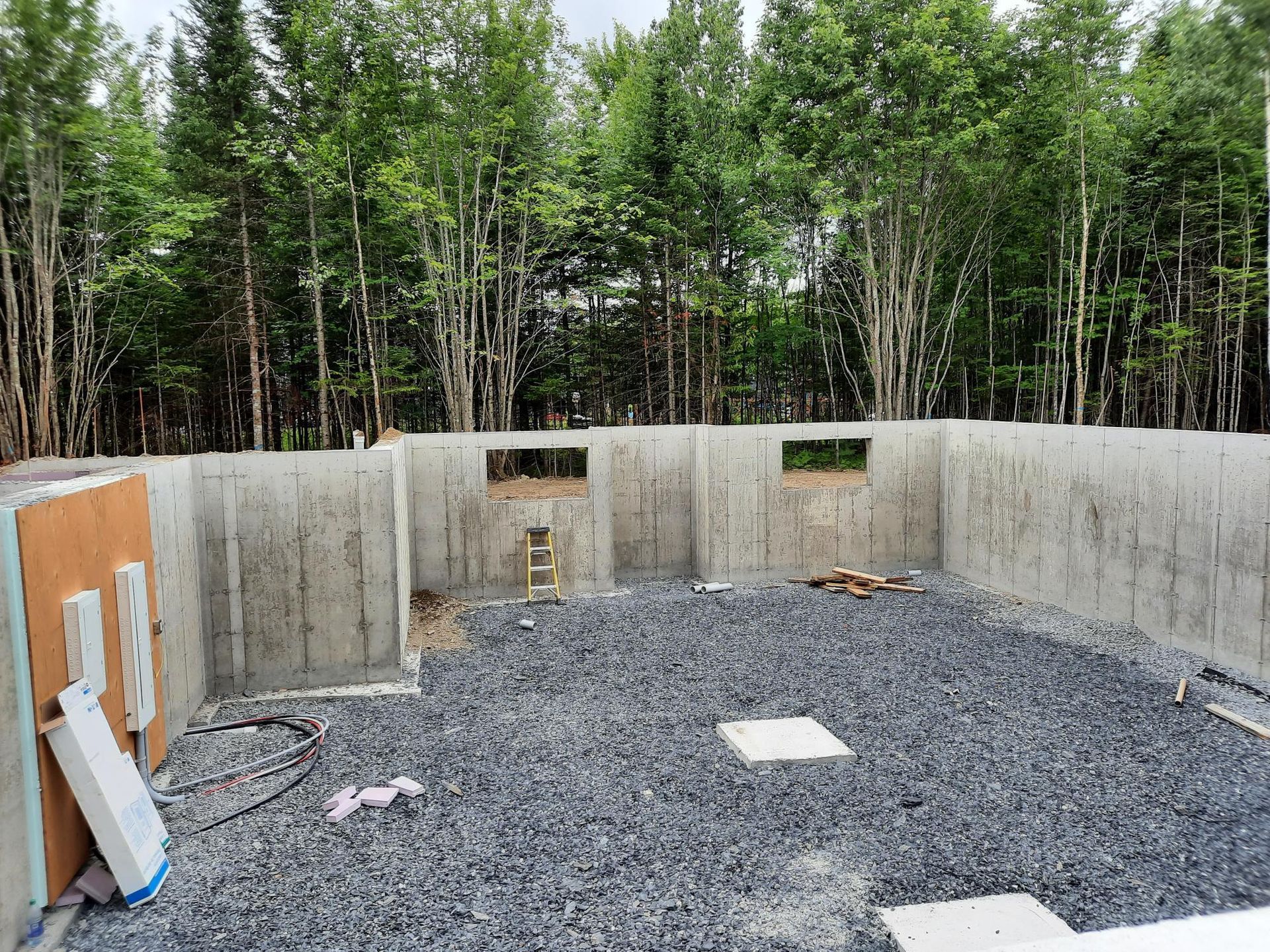 Murs de fondation en béton avec sol en gravier ; fenêtres, arbres en arrière-plan.