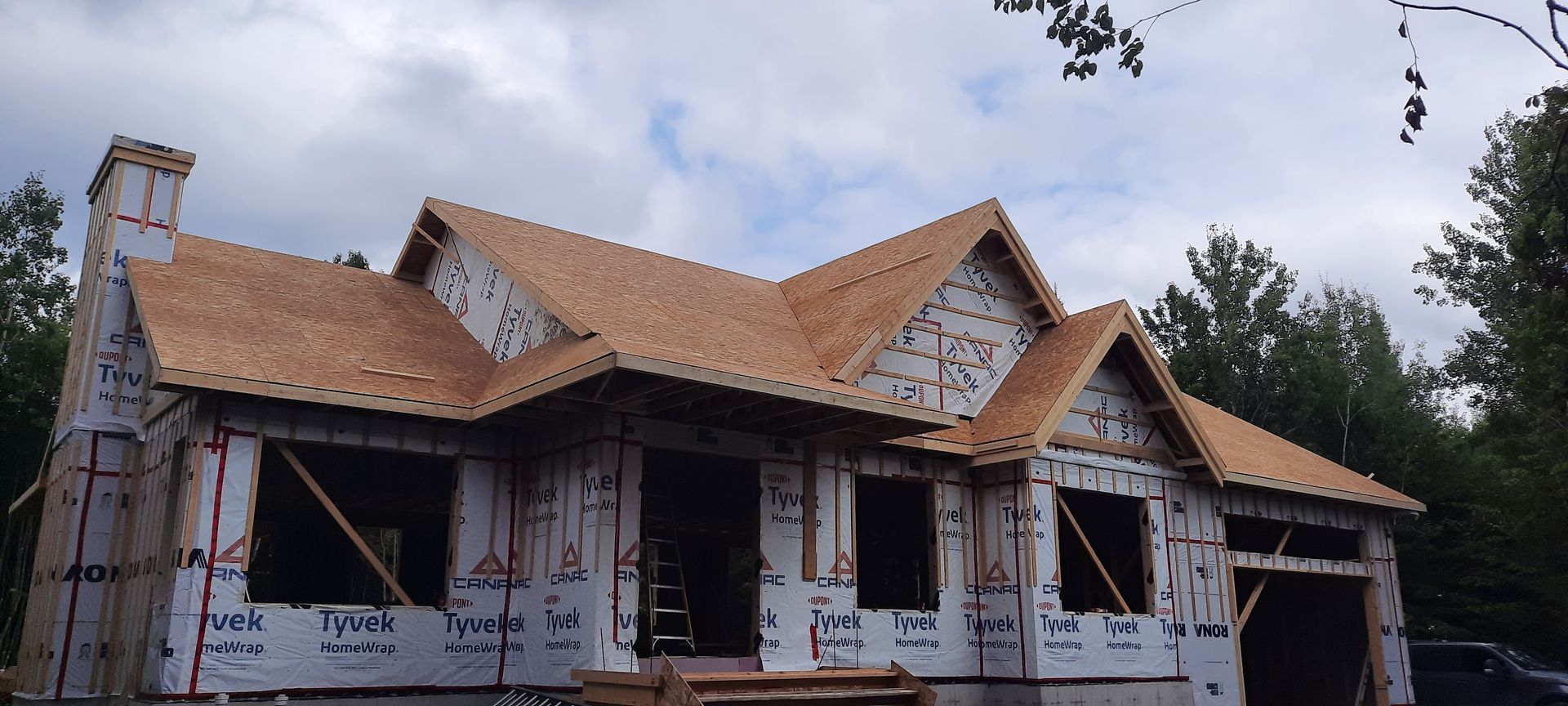 Maison en construction, toiture et charpente visibles, entourée d'arbres et d'un ciel nuageux.