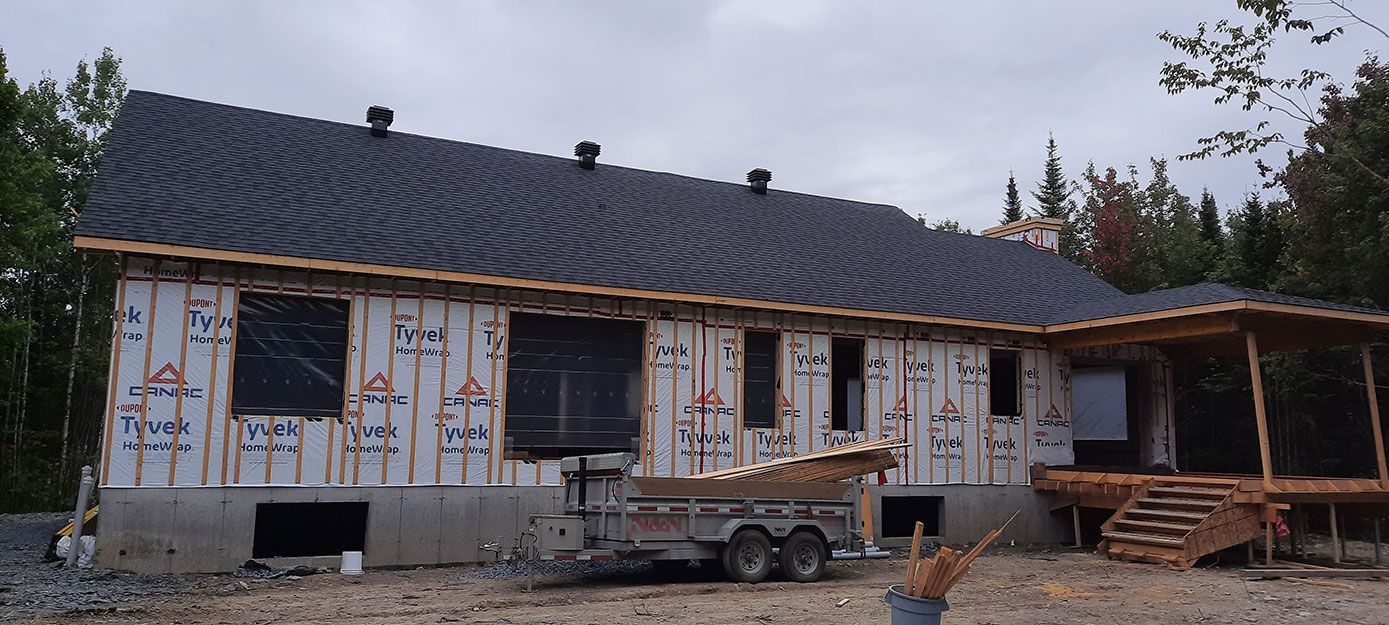Maison en construction avec un toit gris foncé, un bardage inachevé et un porche en bois.