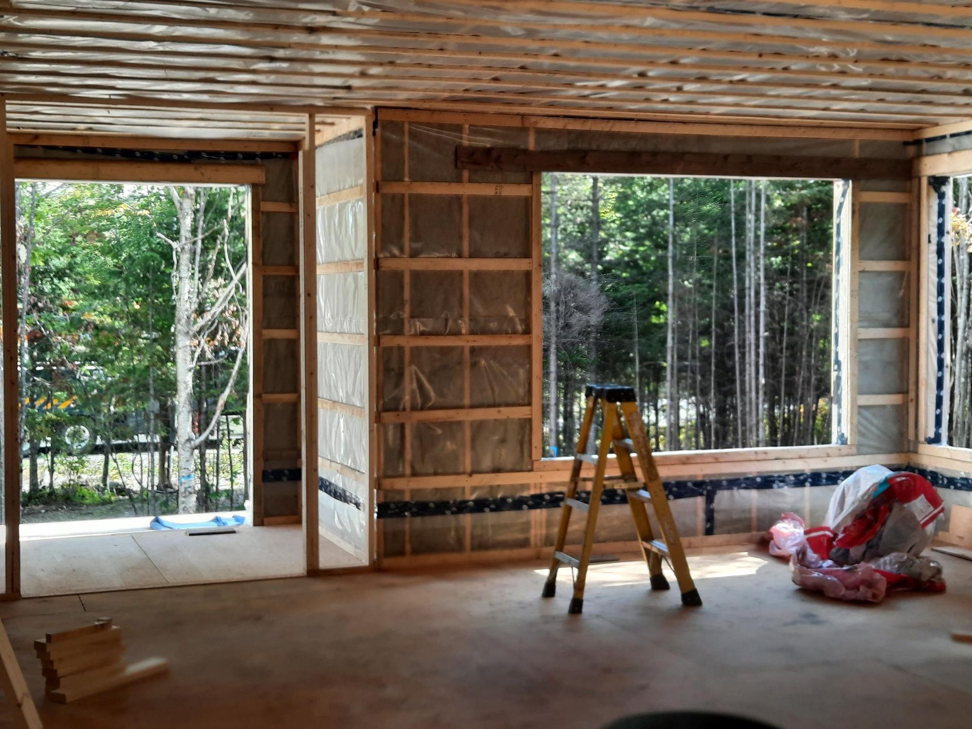 Intérieur d'un bâtiment en construction : charpente en bois isolée, échelle et vue sur les arbres.