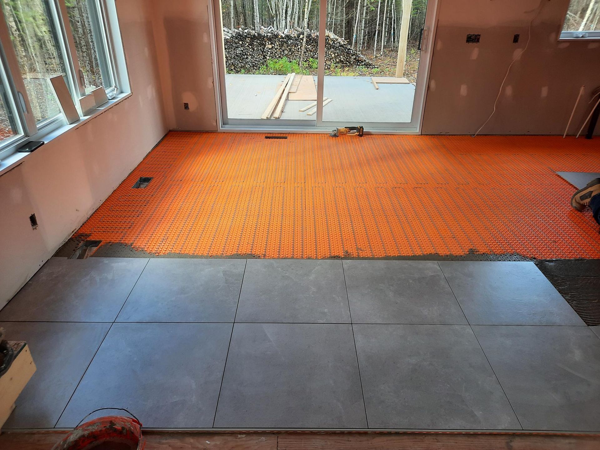 Pièce intérieure avec système de chauffage au sol orange, carrelage gris partiellement posé et porte coulissante en verre.
