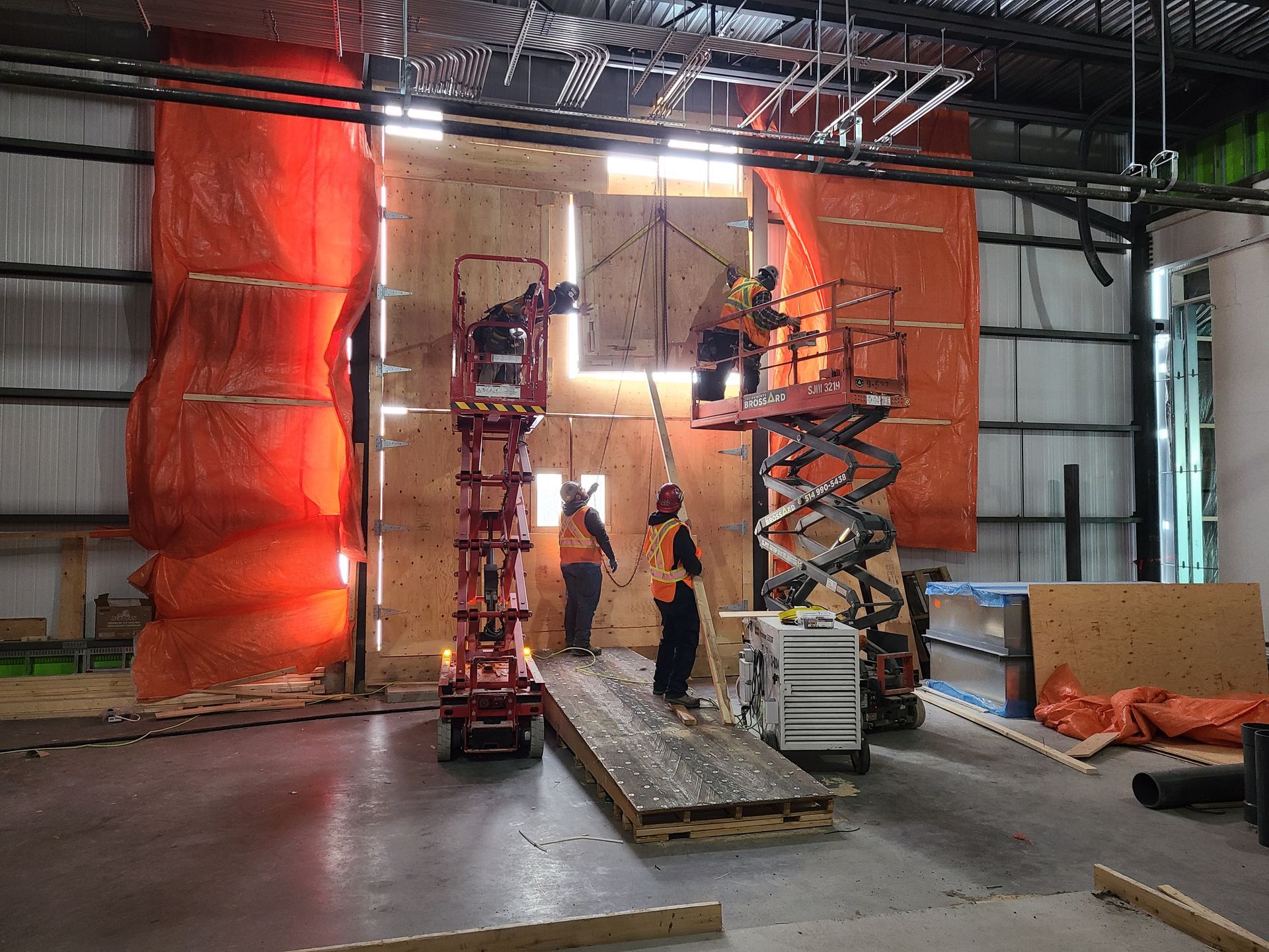 Des ouvriers du bâtiment, sur des nacelles élévatrices, construisent un mur en bois à l'intérieur d'un entrepôt.
