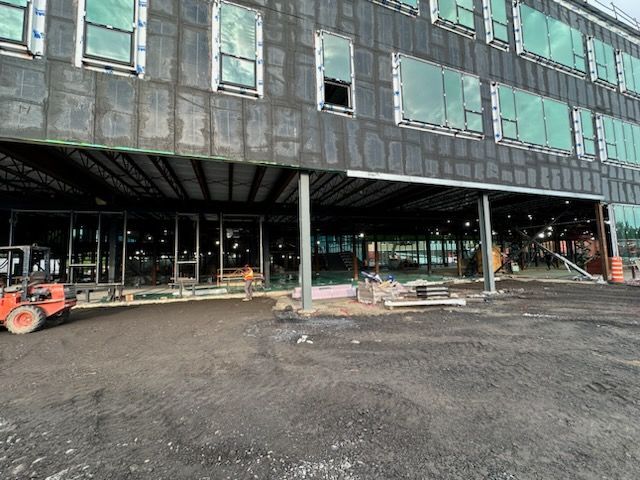 Bâtiment en construction, façade sombre et grandes fenêtres, entrée principale soutenue par des piliers.