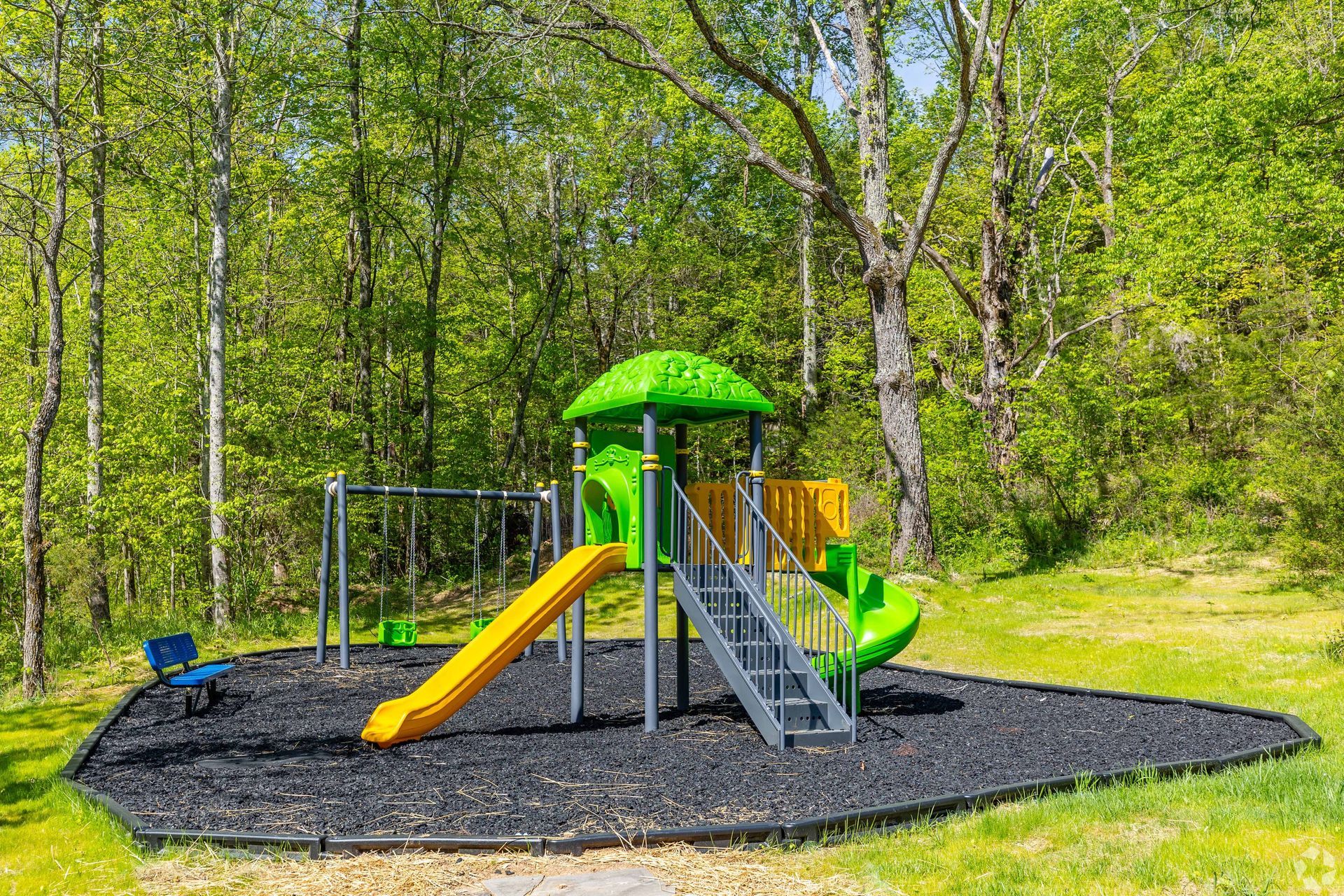 Playground with yellow slide, green roof, and swing set, surrounded by trees and mulch.