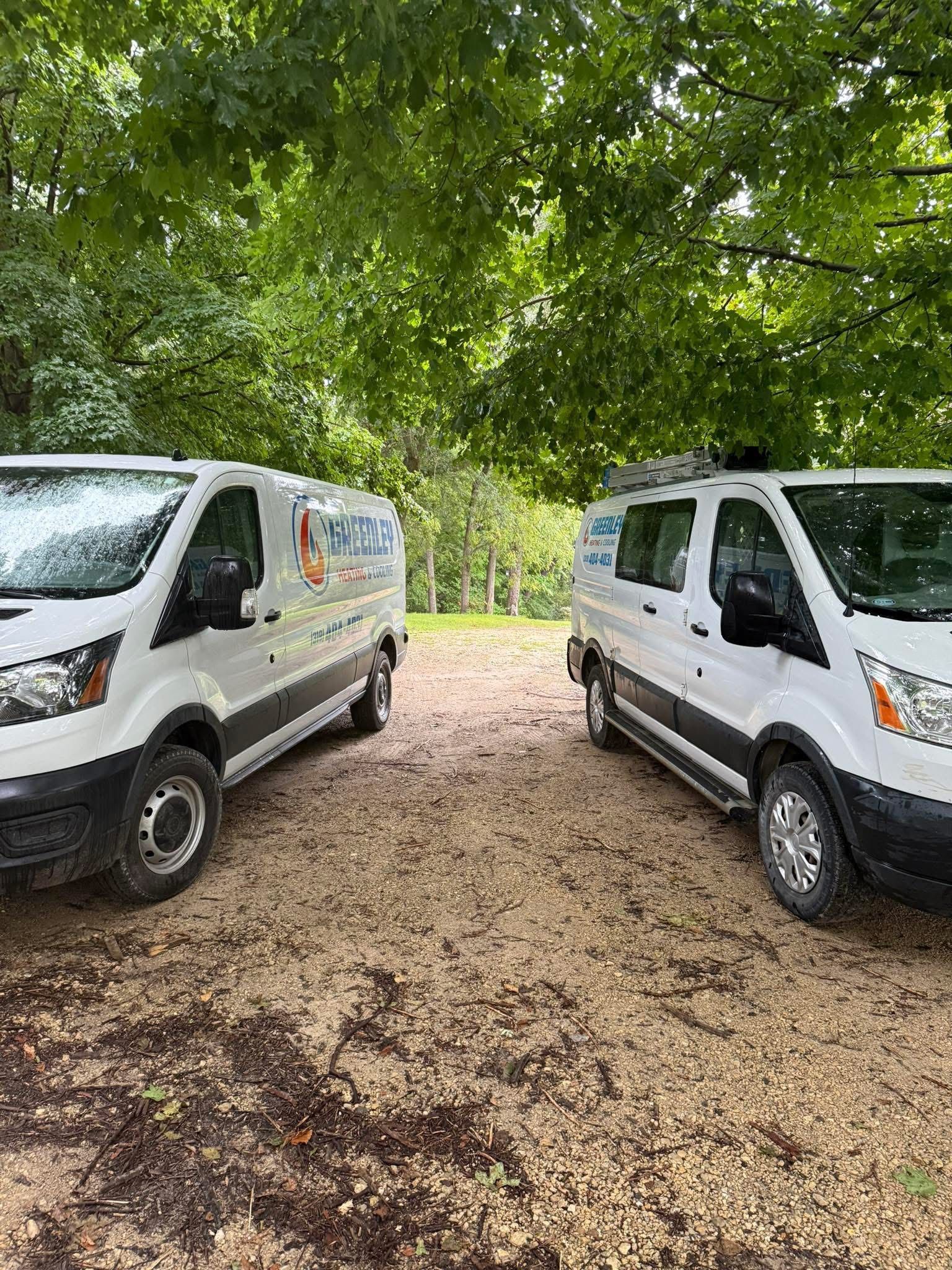 Two white Ford Transit work vans parked side-by-side on a gravel path under trees.