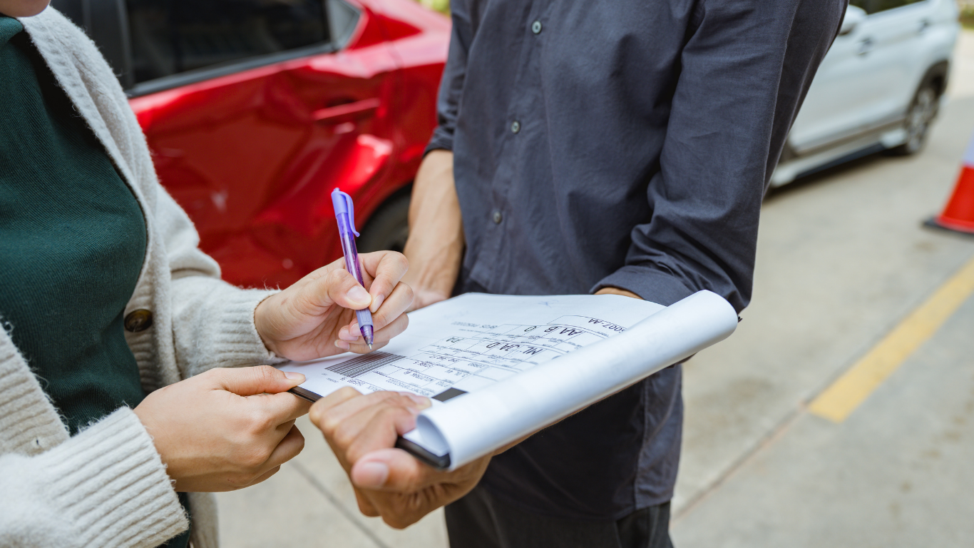 Person signing paperwork held by another person near parked cars.