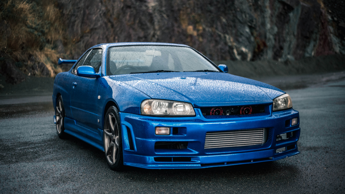 Blue Nissan Skyline R34 car parked on wet asphalt, in front of rocky background.