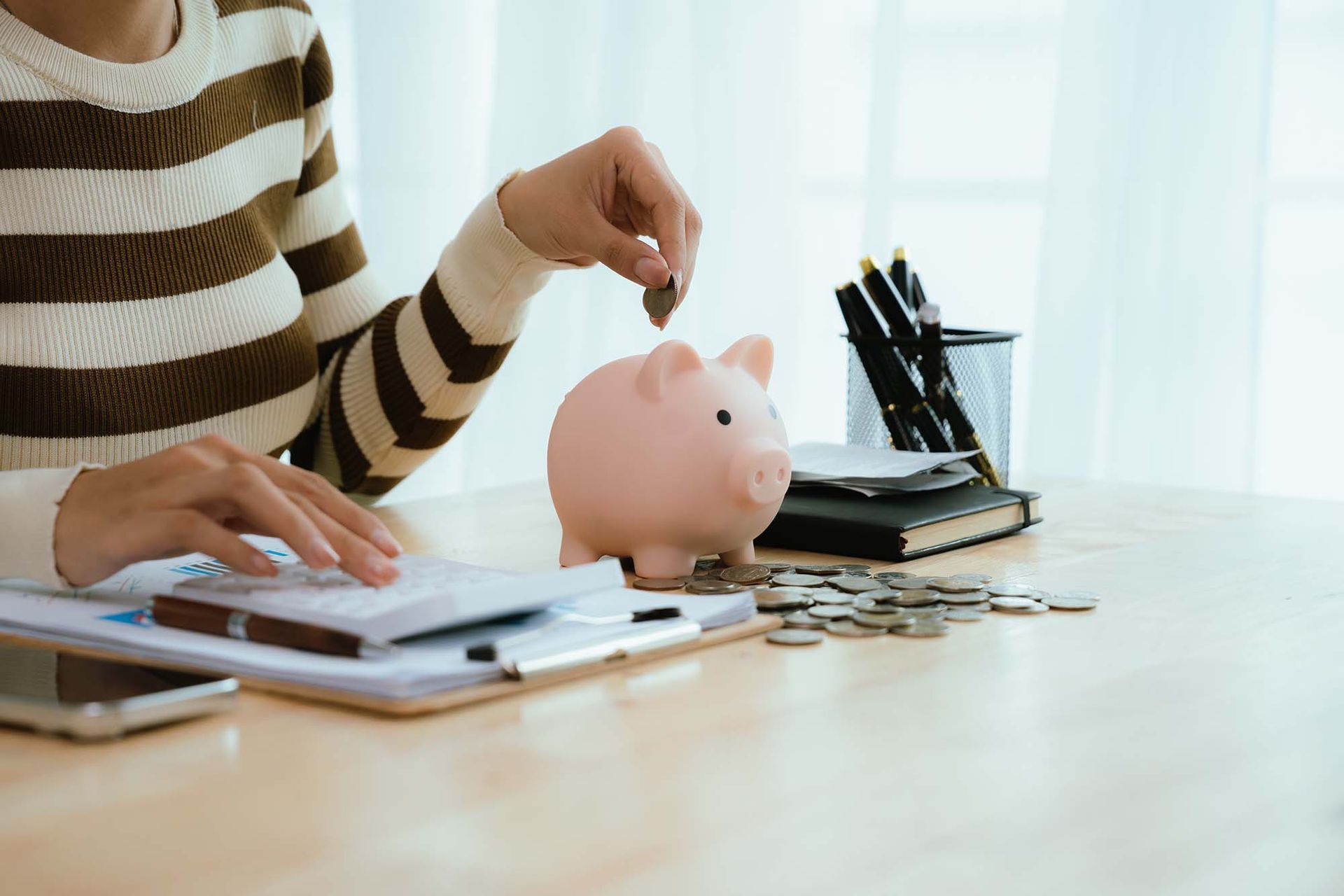 Person placing a coin into a pink piggy bank beside a laptop and scattered coins on a desk