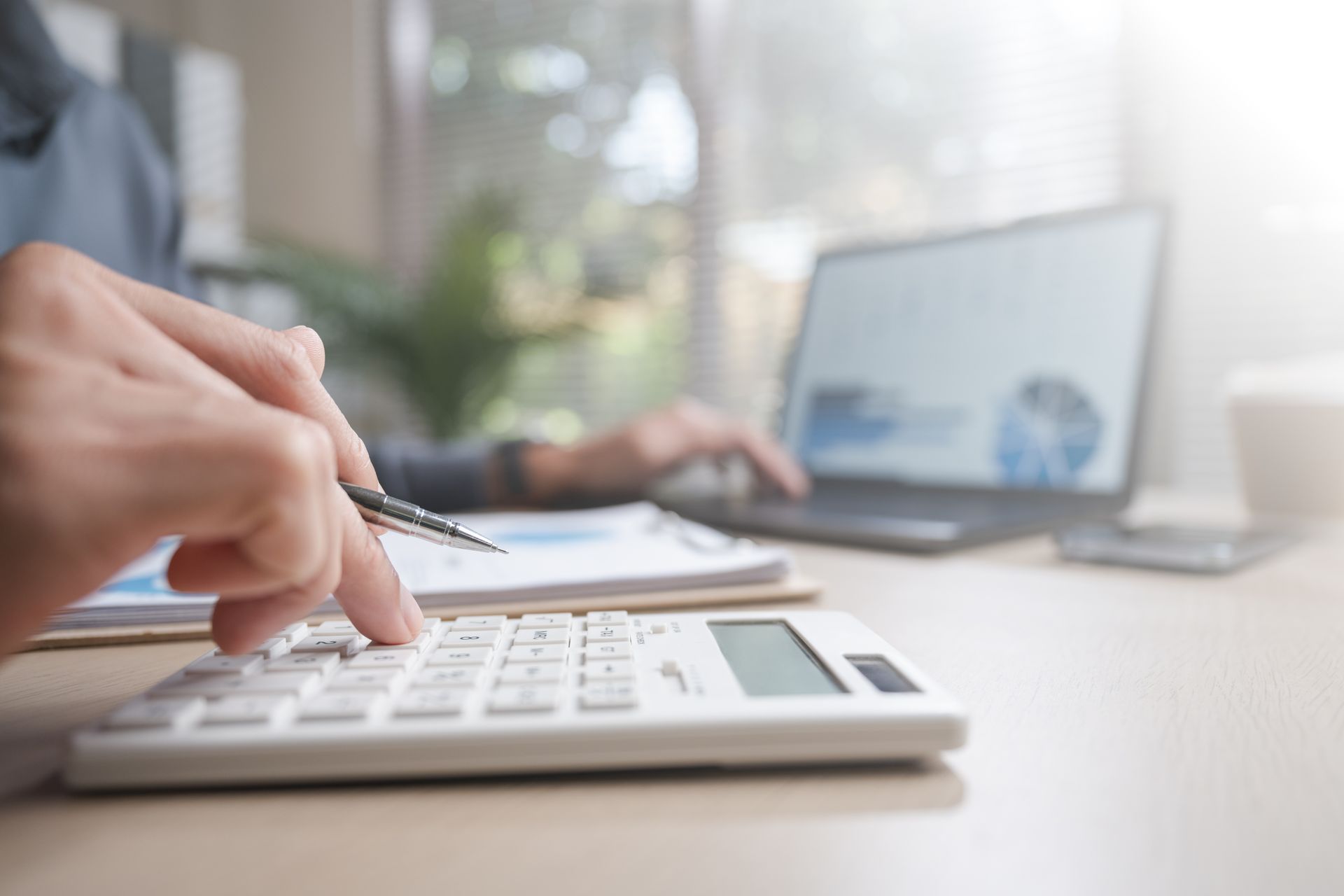 Hand using a calculator beside a laptop in a bright office