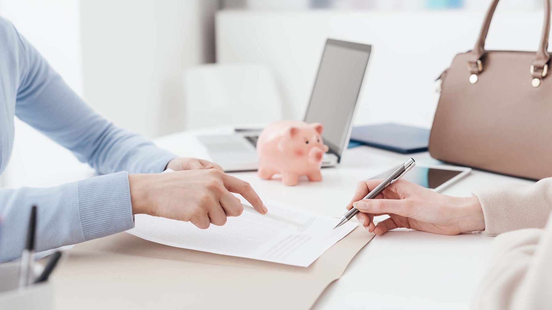 Two people discuss paperwork at a desk with a laptop, piggy bank, and brown handbag.