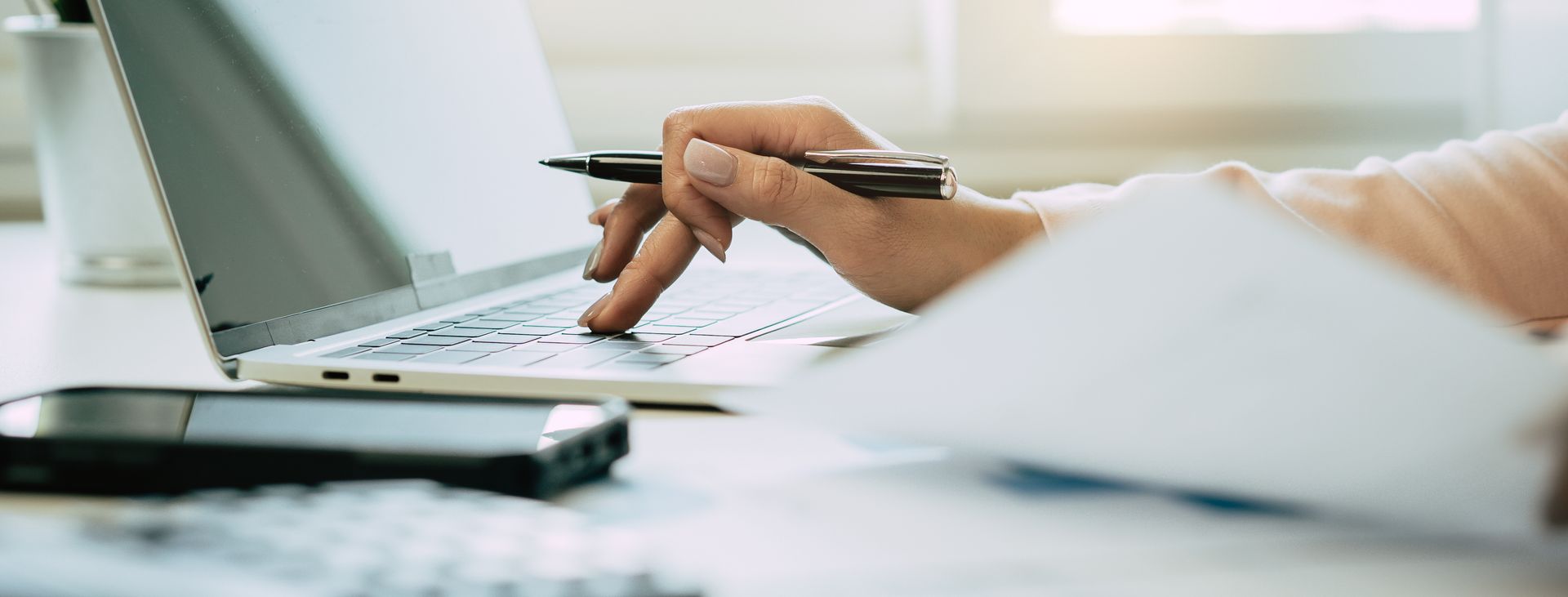 Hand holding a pen over paperwork beside a laptop on a desk